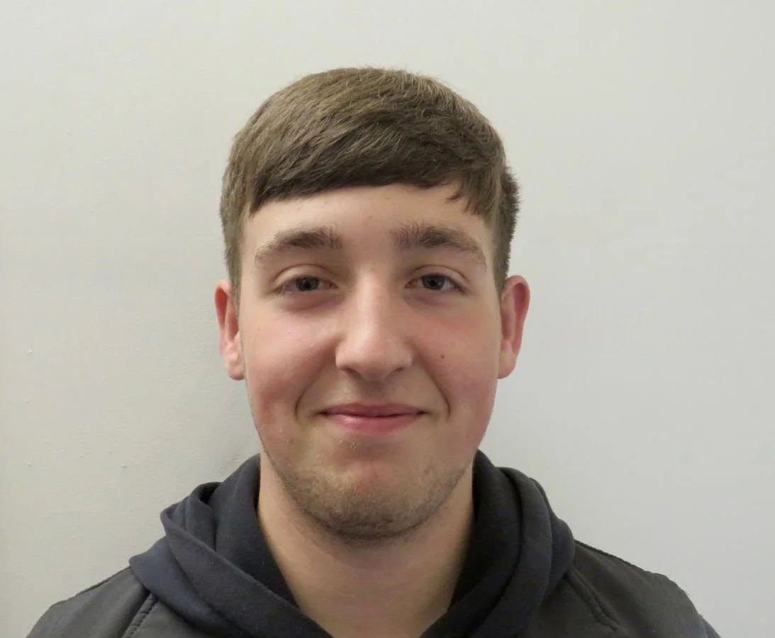 A young man with brown hair and a slight smile, wearing a black hoodie, standing against a plain light-colored wall.