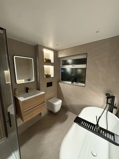Modern bathroom with a wooden vanity, rectangular mirror with LED lighting, wall-mounted toilet, built-in shelf with plants, and a bathtub with a black faucet and a tray. There is a window and neutral-colored walls.