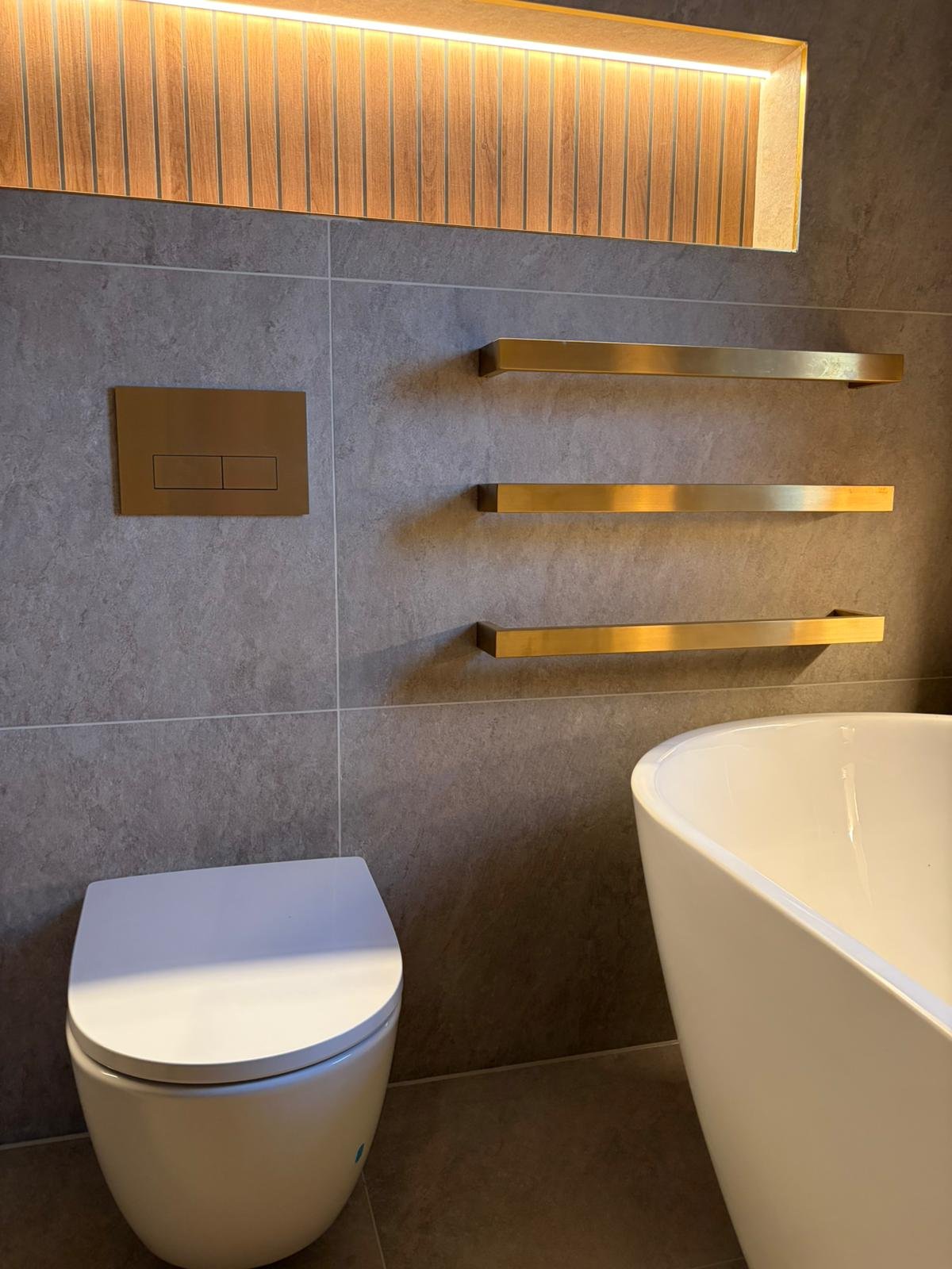 Modern bathroom with gray tiled walls, a wall-mounted gold flush plate, three horizontal gold towel bars, a white oval bathtub, and a portion of a white toilet seat.