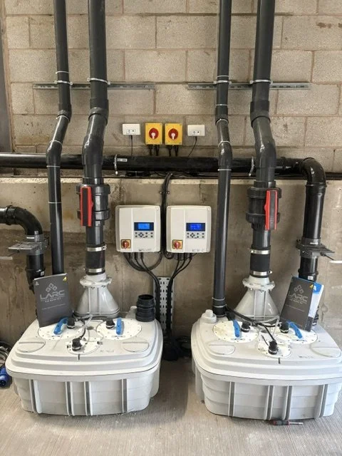 Industrial water pump system with two large white pumps connected to black pipes, control panels, and electrical switches mounted on a concrete wall.