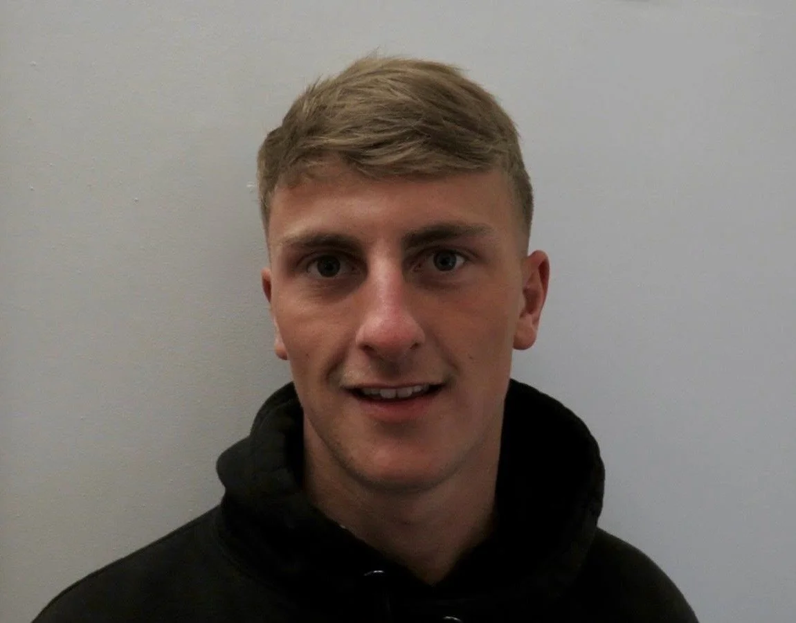 A young man with short blond hair and fair skin, wearing a black hoodie, standing against a plain light gray wall, smiling slightly and looking at the camera.