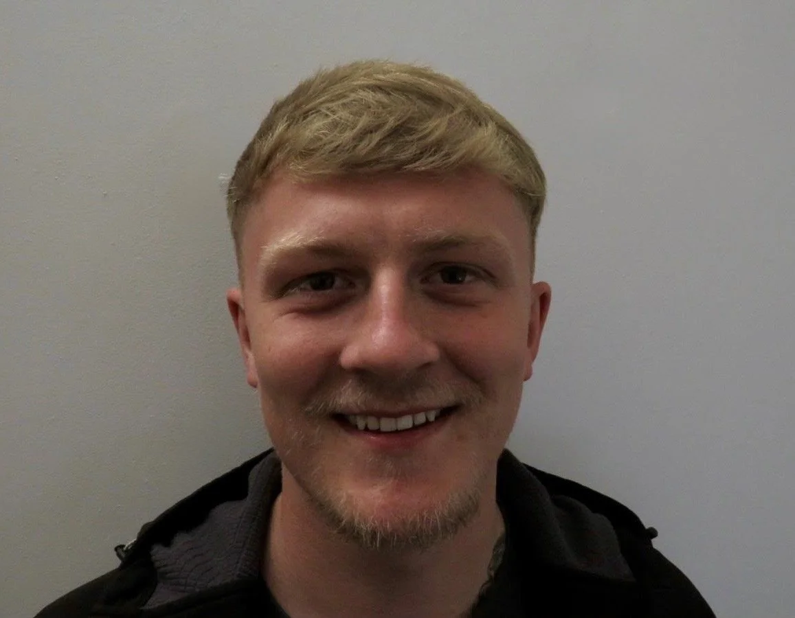 A smiling young man with blonde hair, light facial hair, and wearing a dark jacket standing against a plain gray wall.
