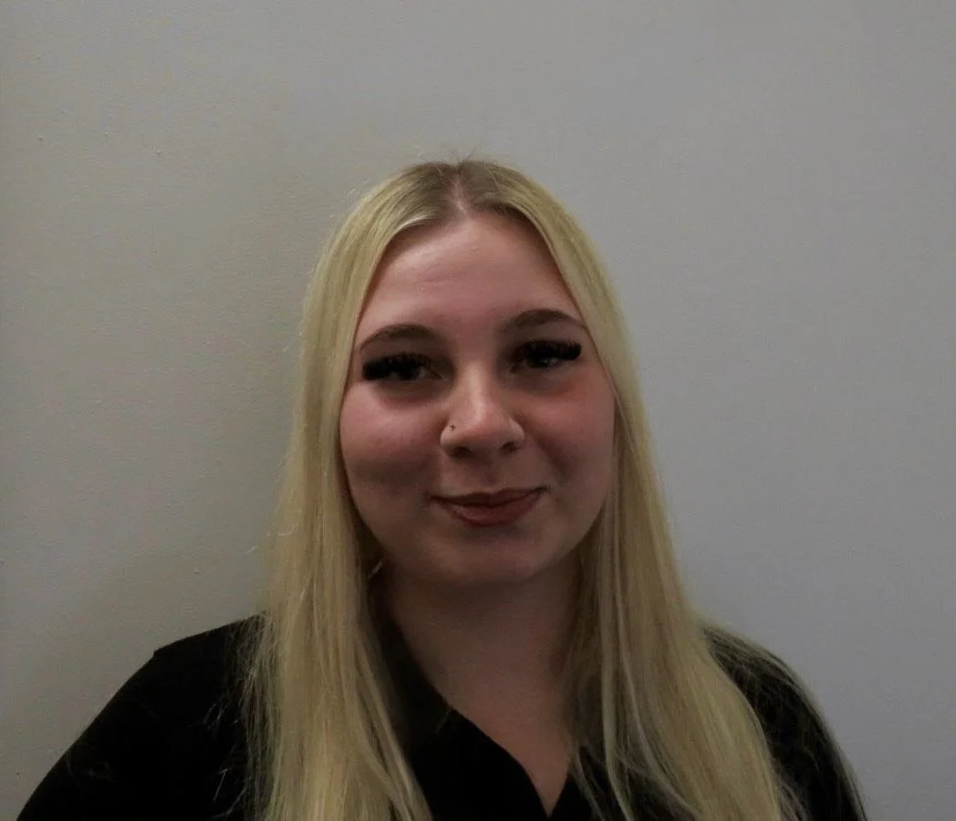 A young blonde woman with a nose piercing smiling in front of a plain wall.
