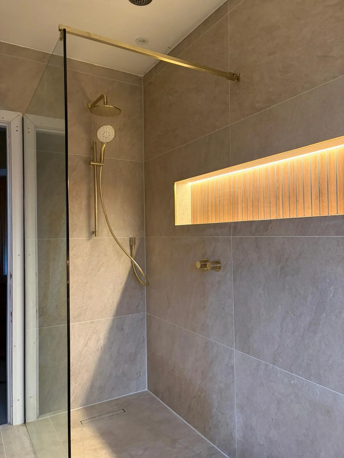 A modern walk-in shower with beige tiled walls and floor, a gold showerhead with a hand-held attachment, a rectangular niche with wood paneling and lighting, and a glass partition.