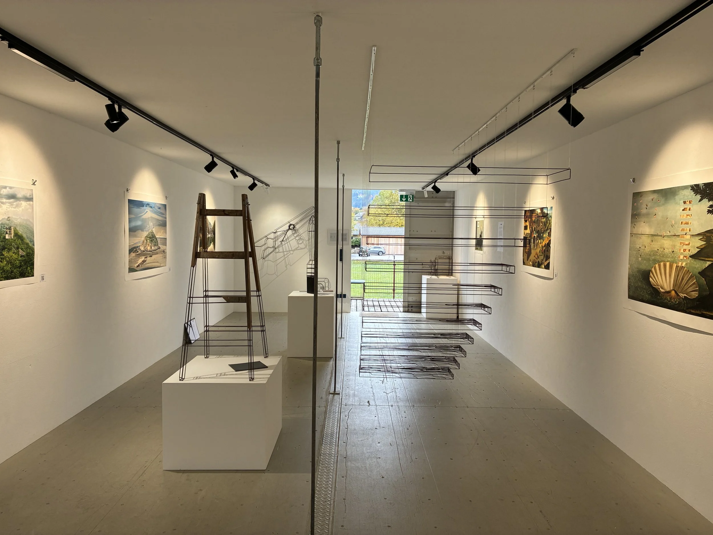 Eine weiße Galerie mit Kunstwerken an den Wänden, einige leere Metallregale und ein Holzleiter, Blick zum Ausgang mit einem wiederholten Fluchtzeichen über der Tür.