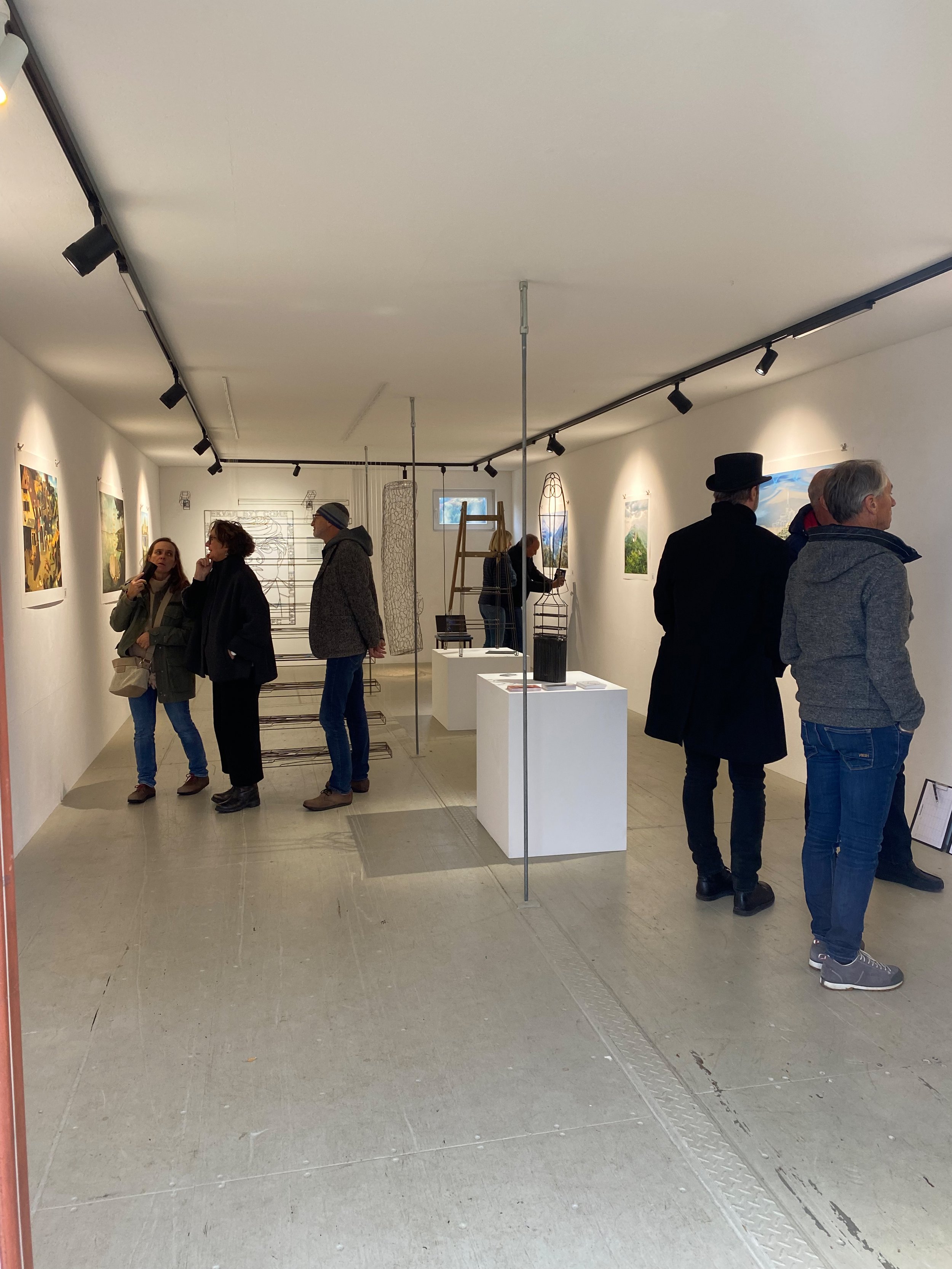 Menschen in einer Galerie betrachten Kunstwerke an den Wänden, einige unterhalten sich, andere schauen sich die Kunst an, es gibt Skulpturen auf Podesten in der Mitte des Raumes.