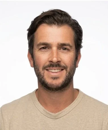 A smiling man with dark hair and a beard, wearing a beige T-shirt, against a plain white background.