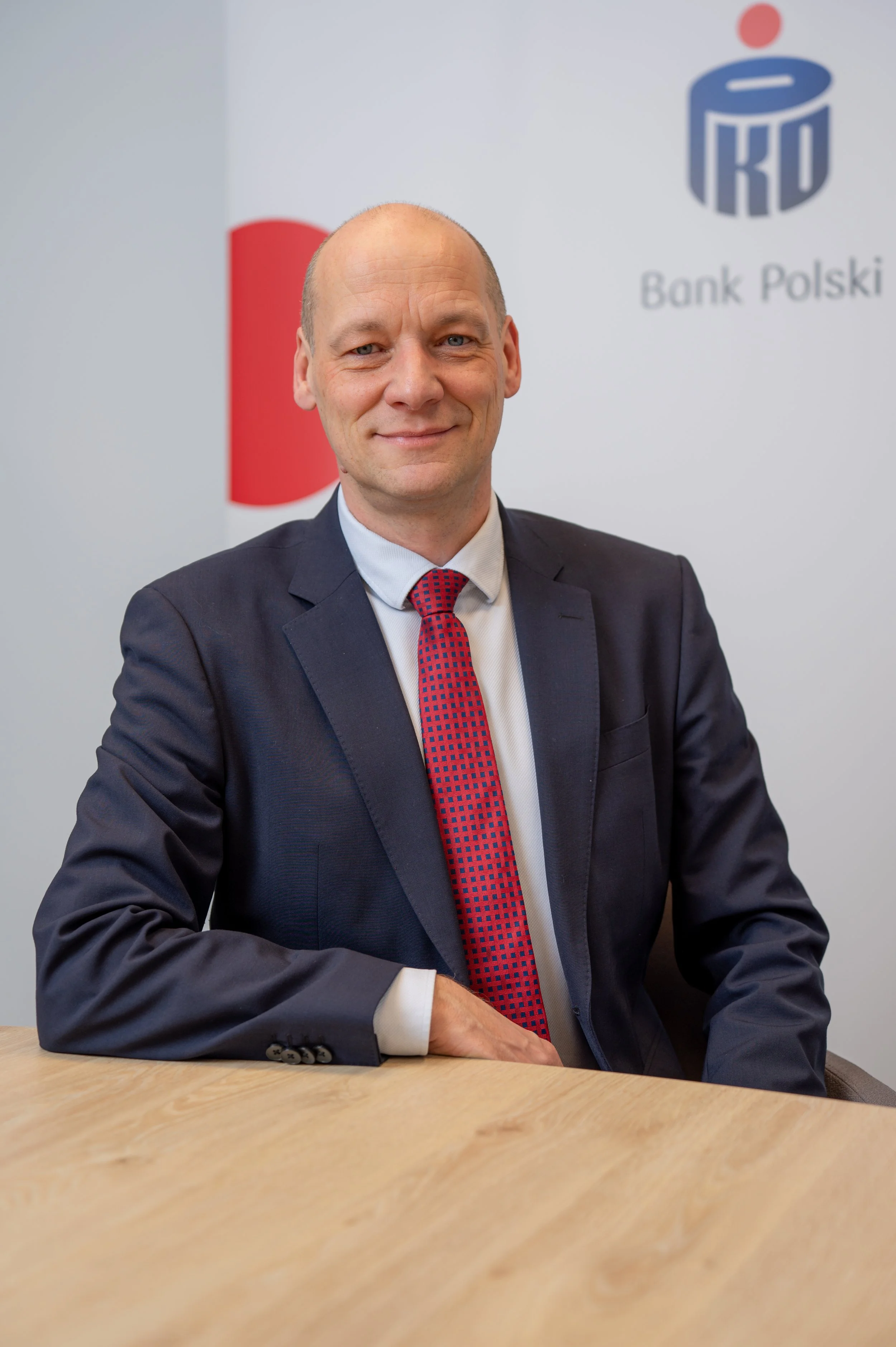 A smiling man in a dark suit, white shirt, and red patterned tie sitting at a table in front of a white background with the logo of Bank Polski and a red and blue emblem.