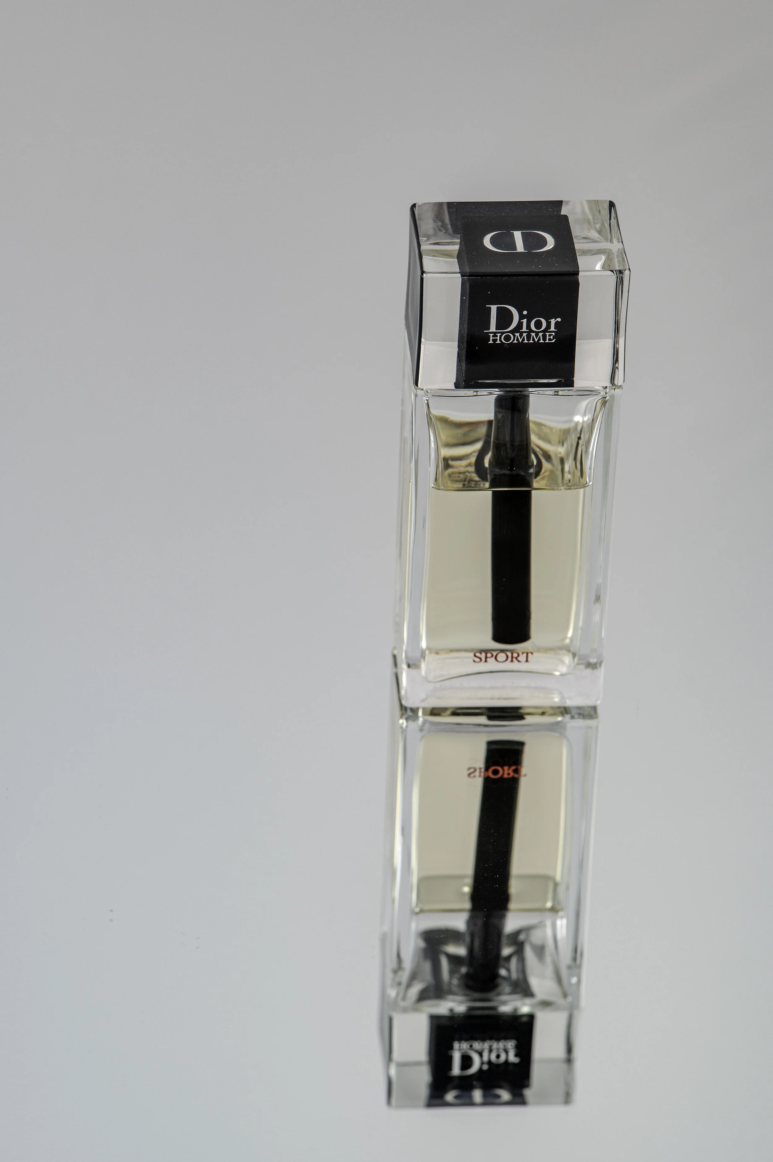 A clear glass rectangular perfume bottle with a black cap, labeled Dior Homme, with the word 'SPORT' in red near the base.