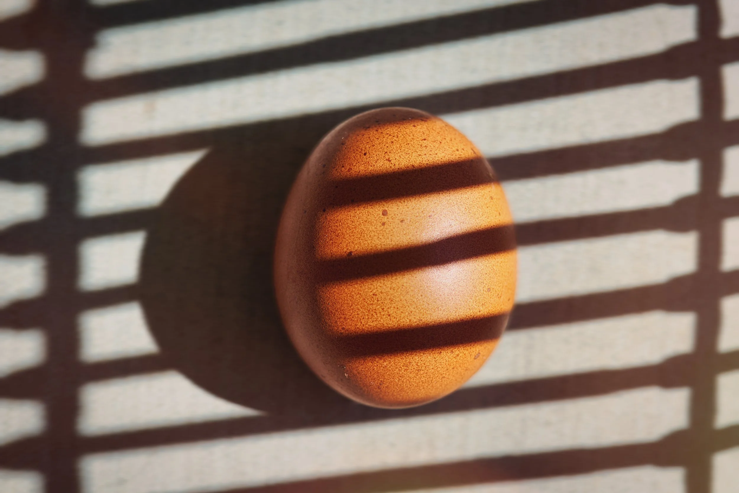 A close-up of an orange egg with a shadow cast on it through a slatted surface, creating horizontal striped shadows on the egg.