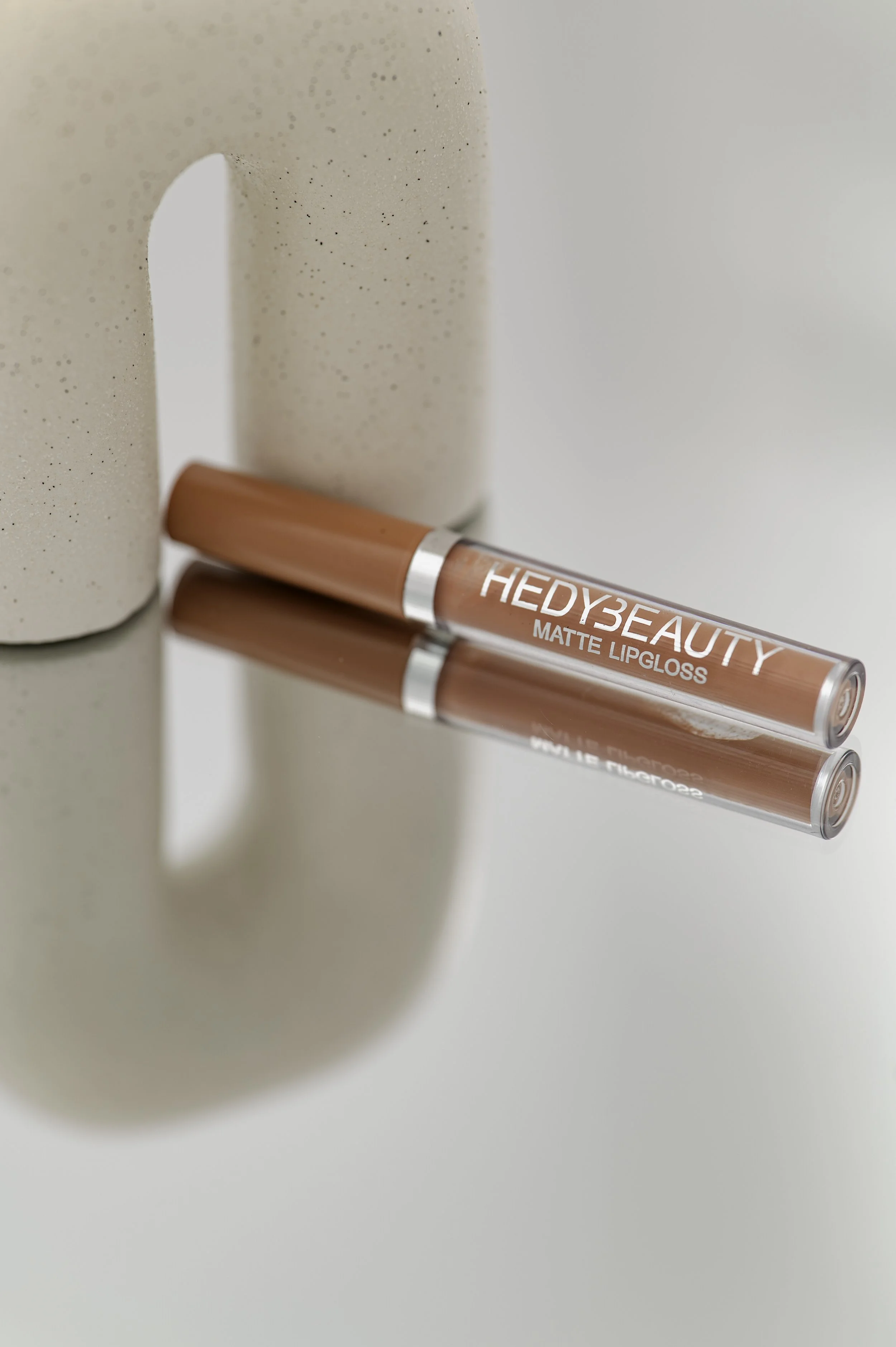 Close-up of a beige lipstick tube labeled "HEDY3BEAUTY MATTE LIPGLOSS" lying on a mirrored surface with a beige speckled ceramic mug beside it.