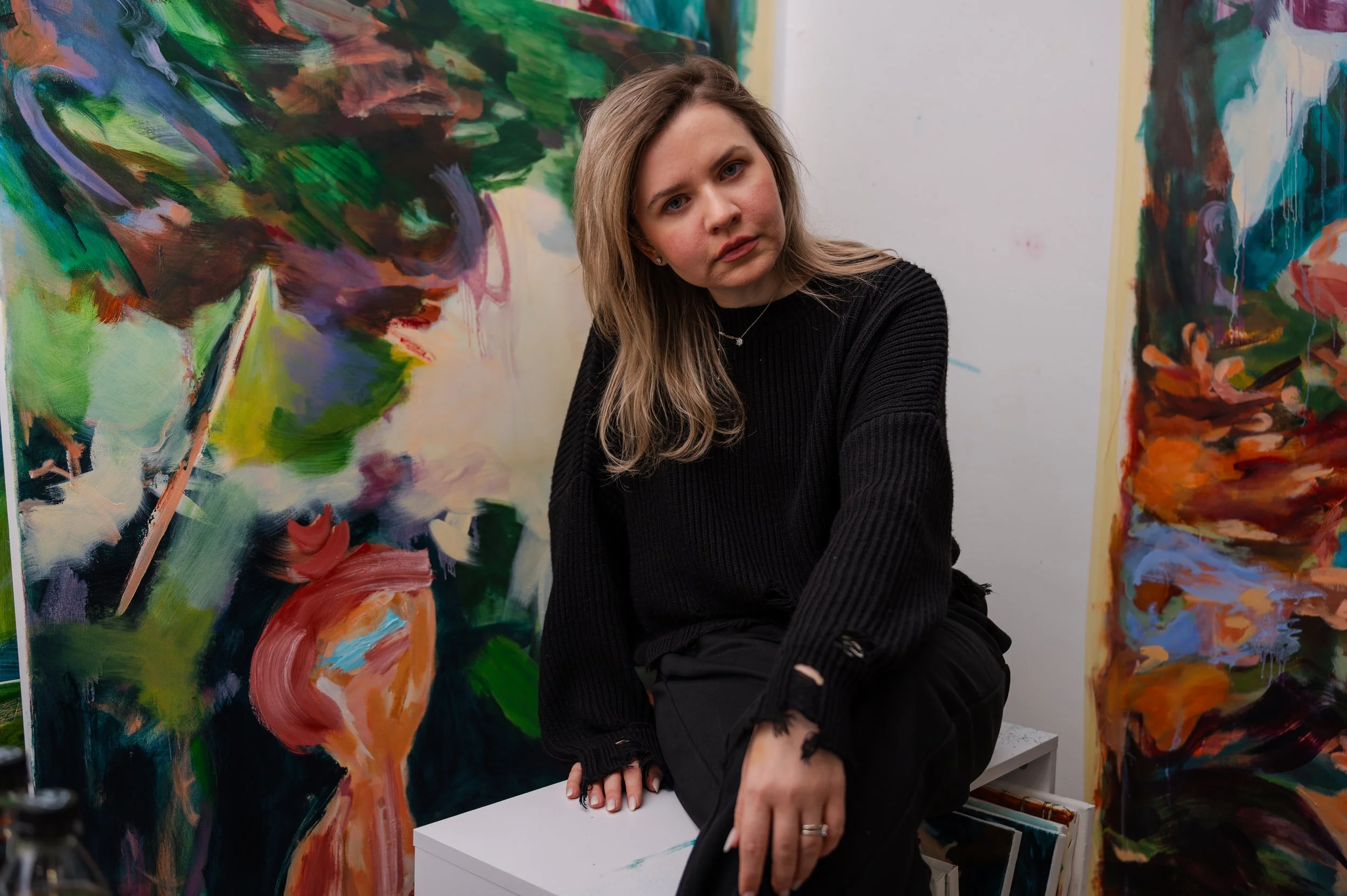 A woman with brown hair and wearing a black sweater is sitting at an art exhibit. She is surrounded by colorful, abstract paintings.