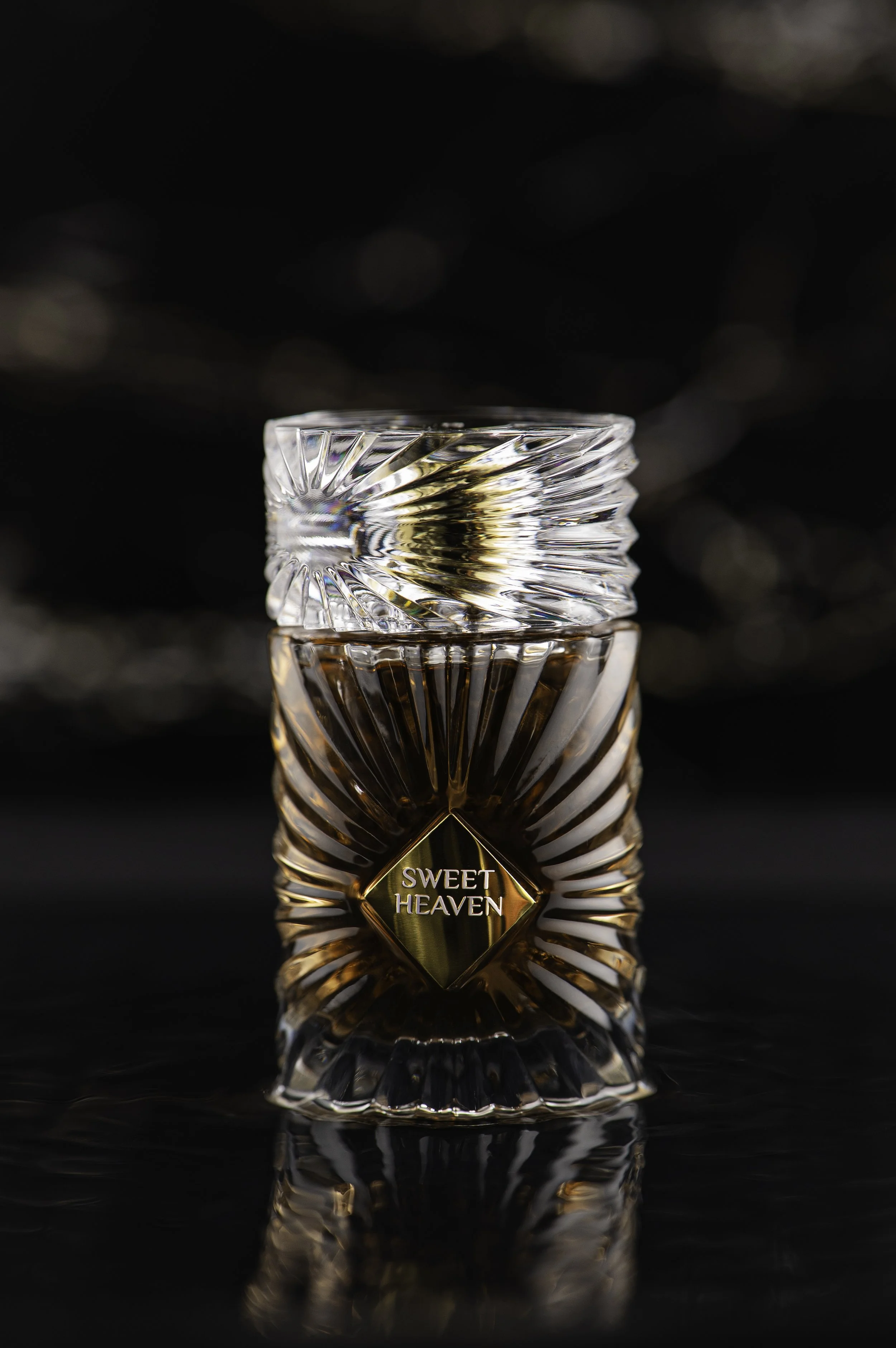 a decorative glass container with a gold diamond-shaped label reading 'SWEET HEAVEN' at the bottom, placed on a reflective black surface with a dark, blurry background