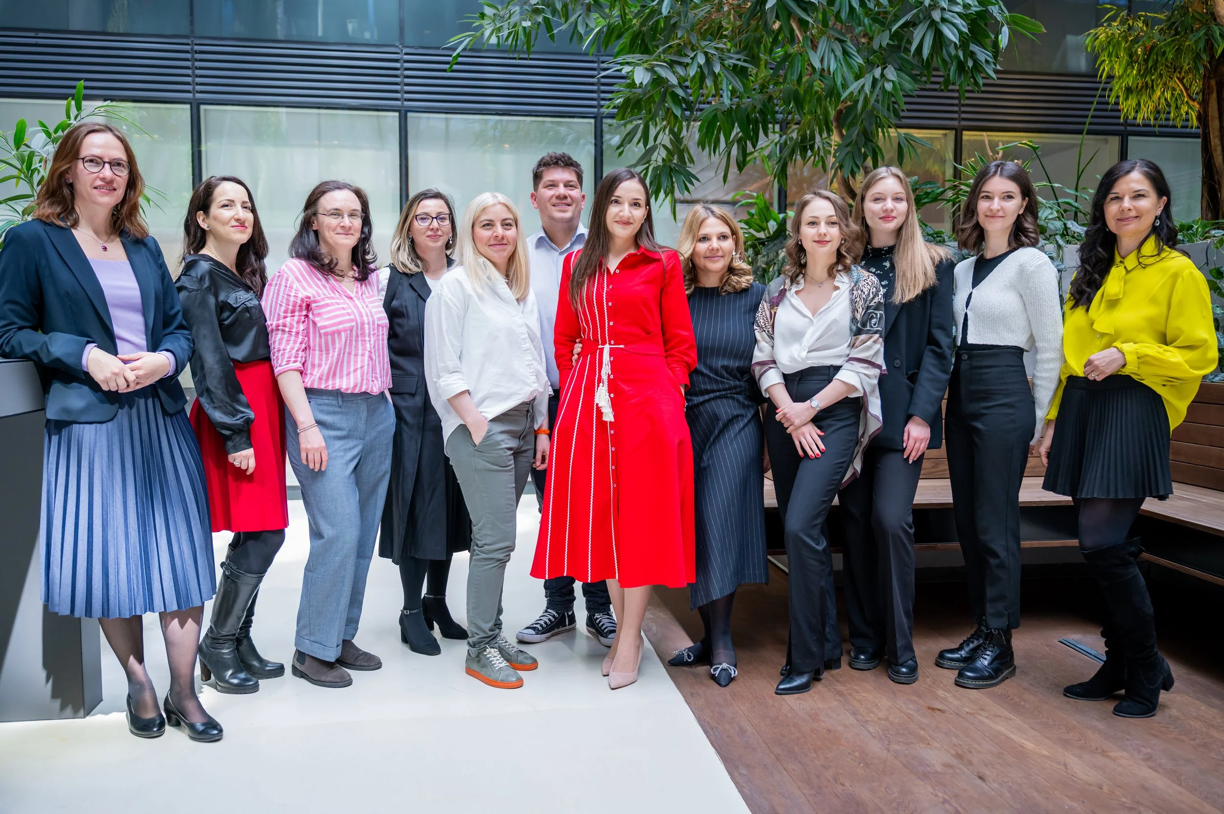 Group of 12 diverse people standing together indoors, posing for a photo with plants and modern building background.