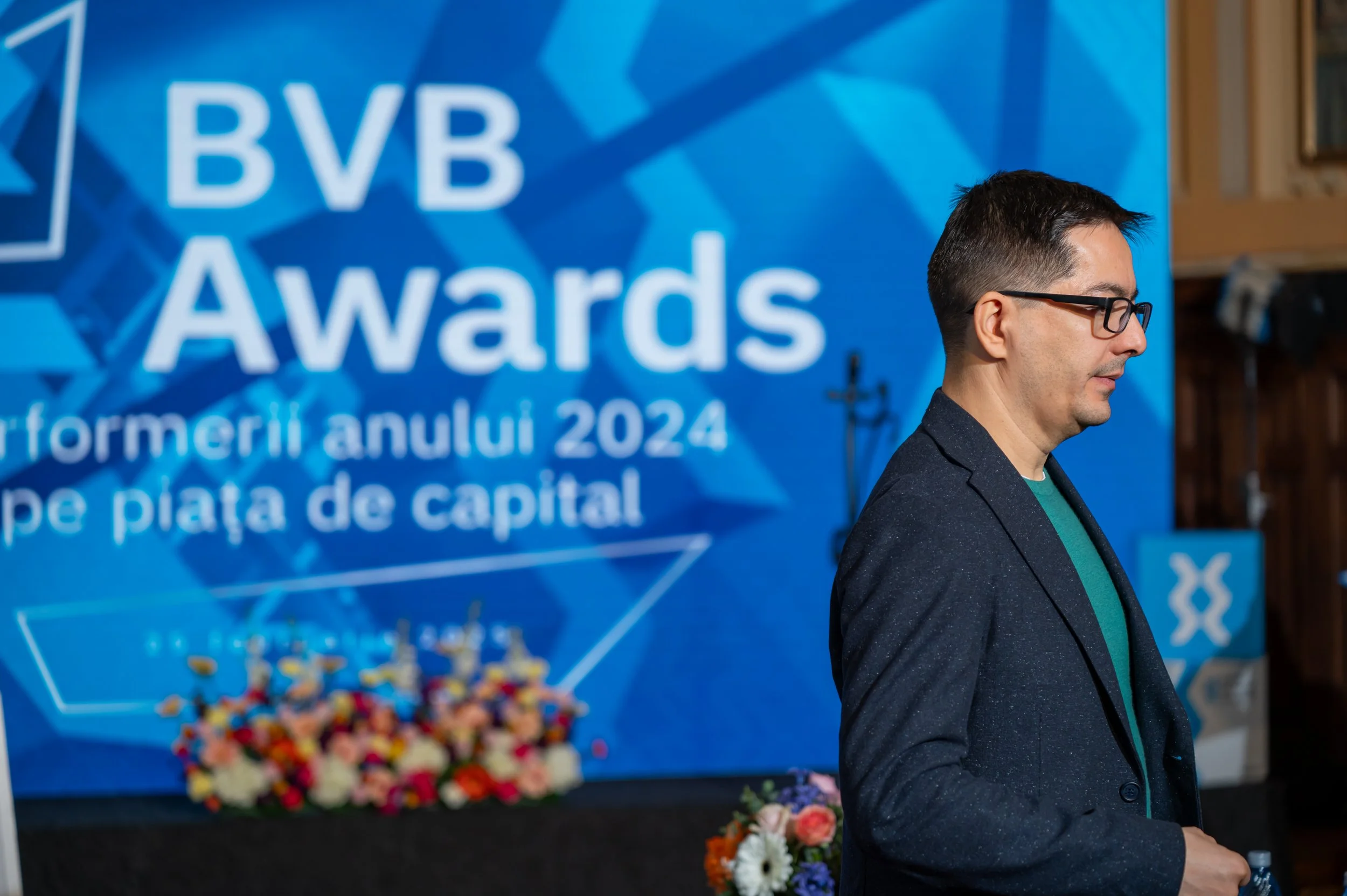 A man with dark hair, wearing glasses, a navy blazer, and a teal shirt, standing in front of a blue sign that reads 'BBB Awards' with additional text in a foreign language, and a colorful floral arrangement in the foreground.