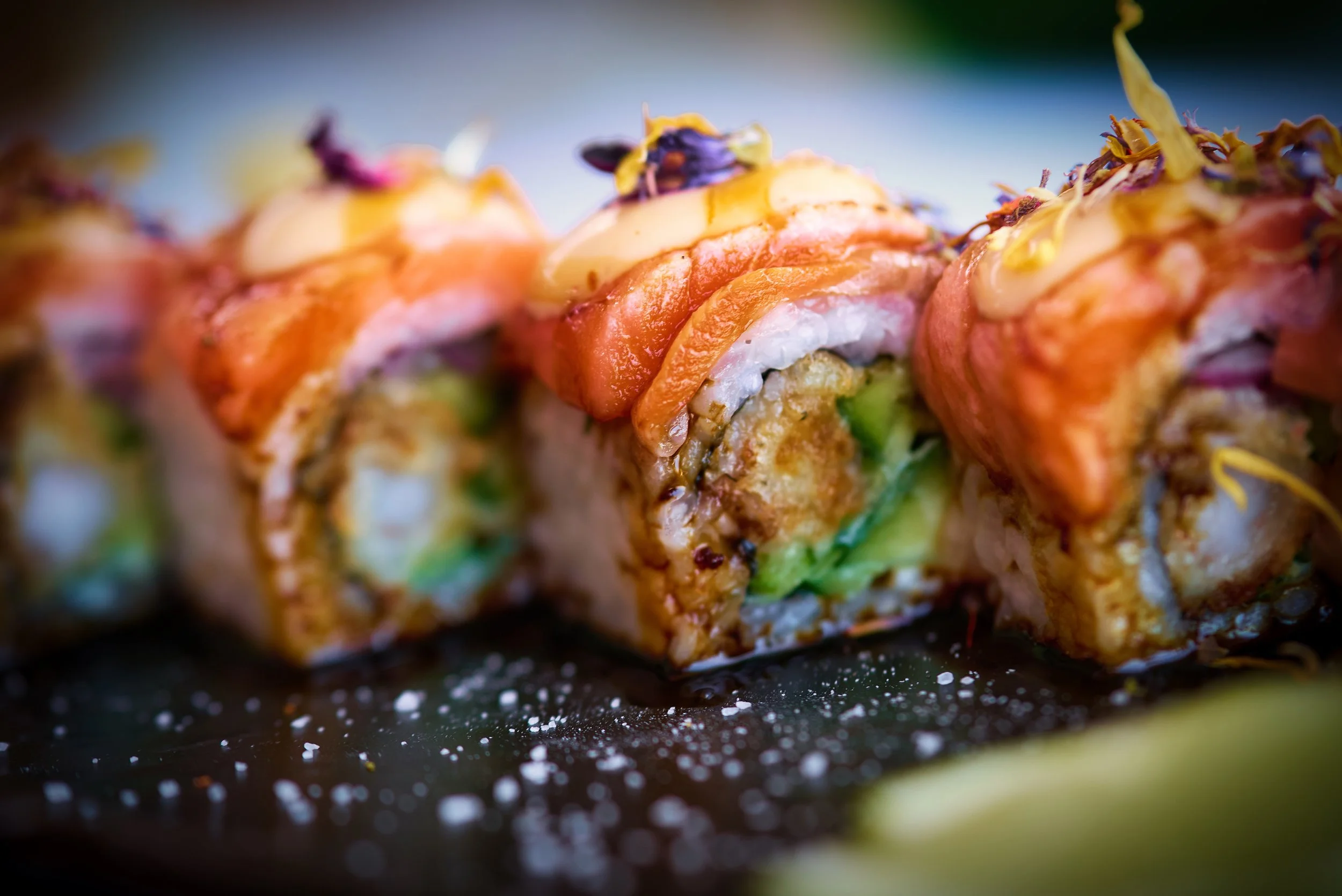 Close-up of colorful sushi rolls garnished with edible flowers and sesame seeds, placed on a dark surface.