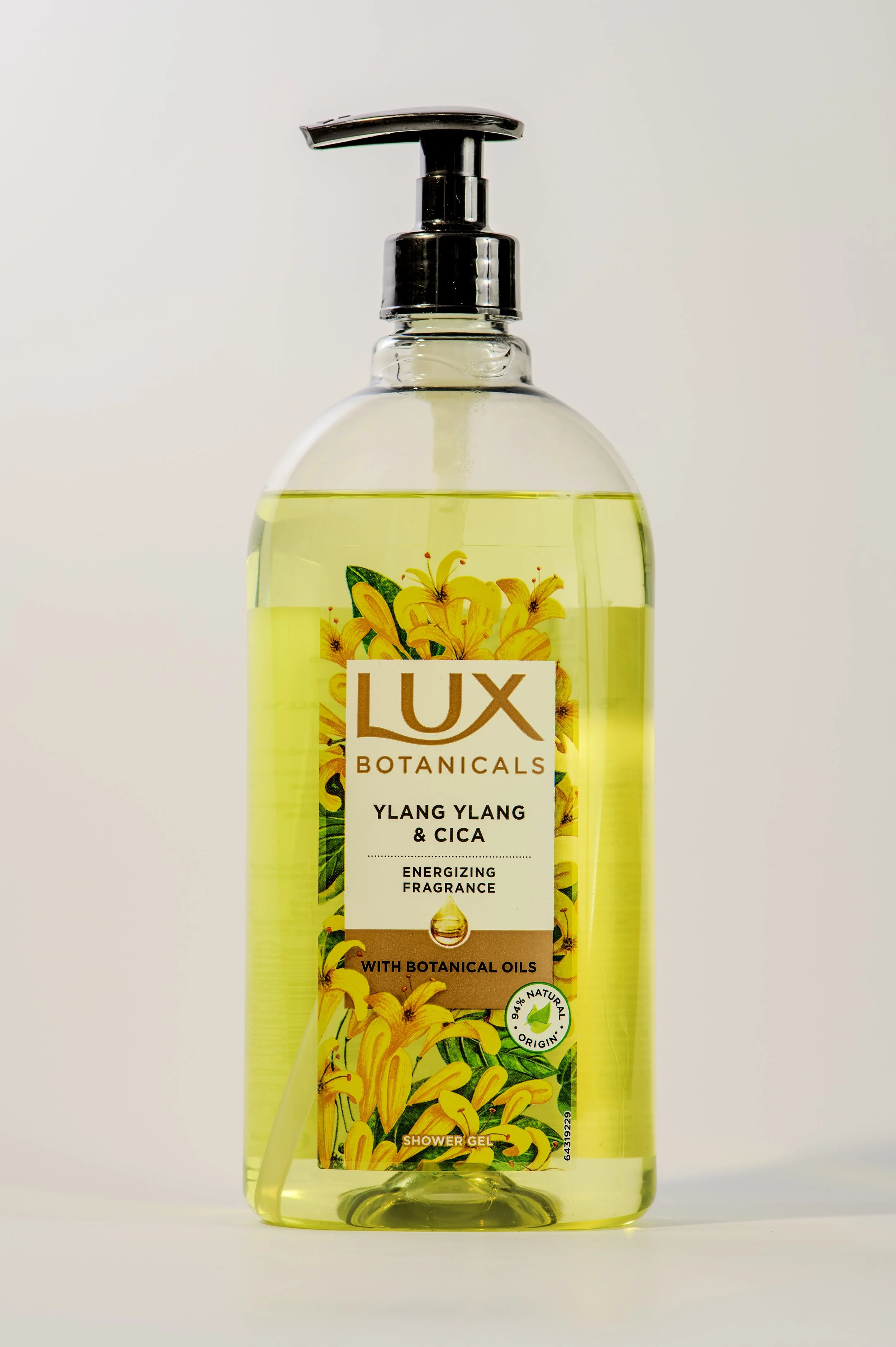 A clear plastic bottle of Lux Botanicals shower gel with a pump dispenser. The label features yellow lilies and green leaves, and indicates it has energizing fragrance with ylang ylang and cica, containing botanical oils, and is made with 94% natural