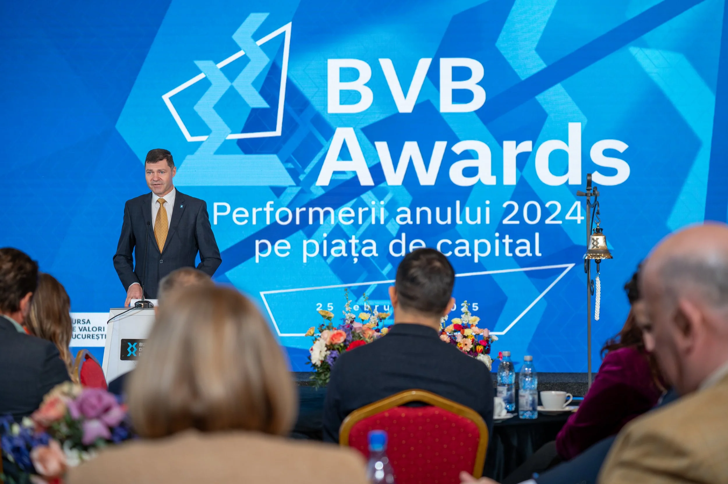 A man in a suit and yellow tie speaking at a conference or awards ceremony. Behind him is a large screen displaying the words 'BVB Awards' and 'Performeri anului 2024 pe piața de capital.' Audience members are seated facing the stage. There are flowe