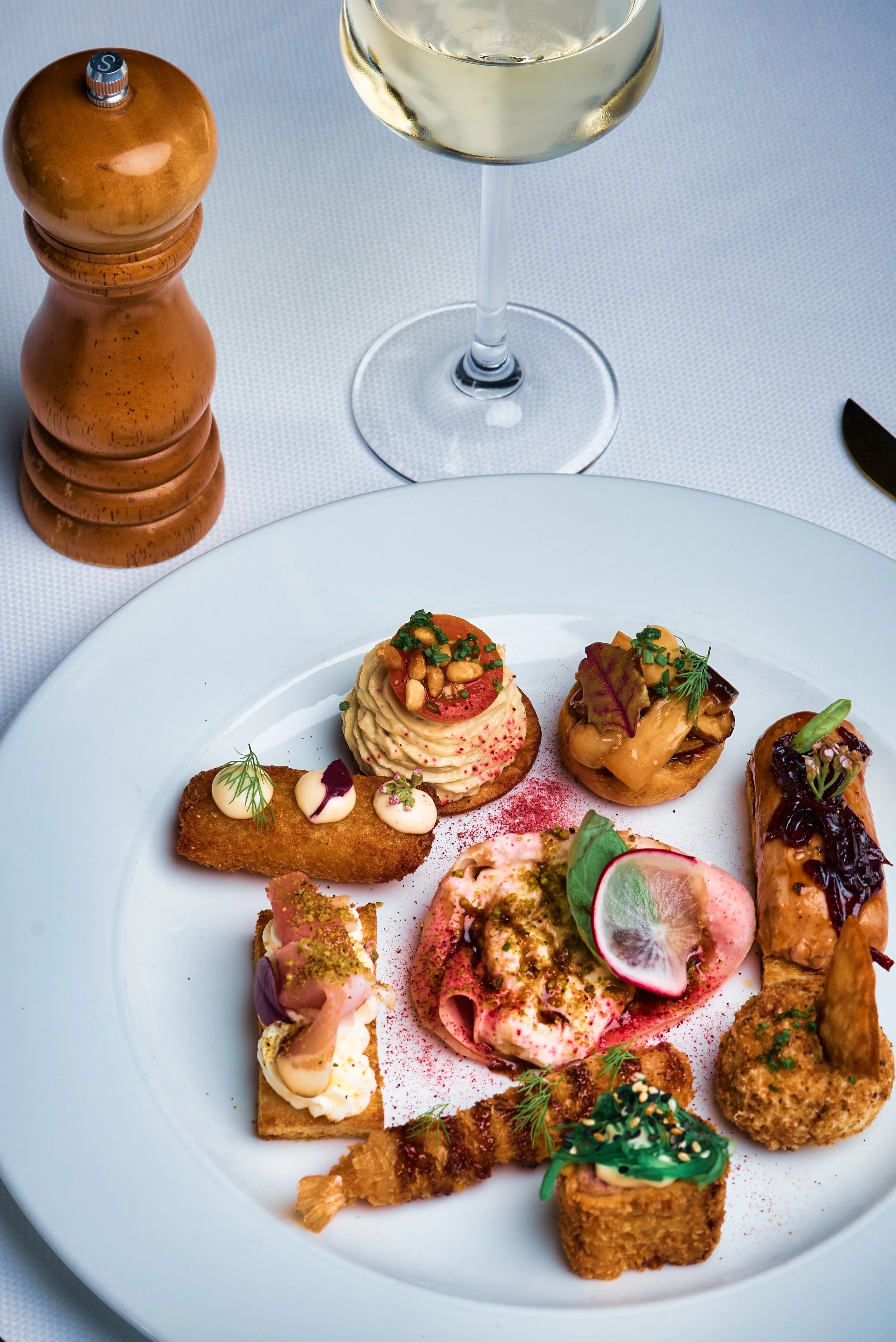 Assorted gourmet appetizers, including a small burger with toppings, sliced radishes, a fried item with green sauce, and finger foods with garnishes, on a white plate at a dining table with a glass of white wine and a salt grinder.