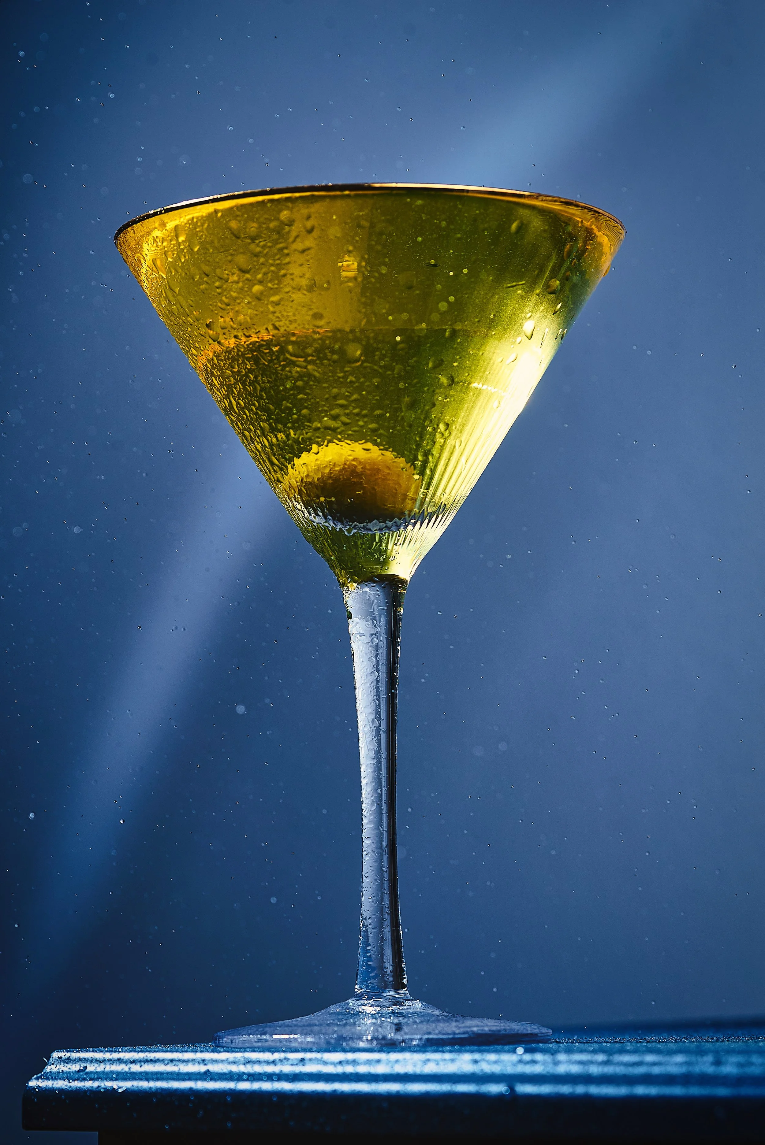 Close-up of a champagne glass with droplets of condensation, filled with a green liquid and a lime wedge, against a blue background.