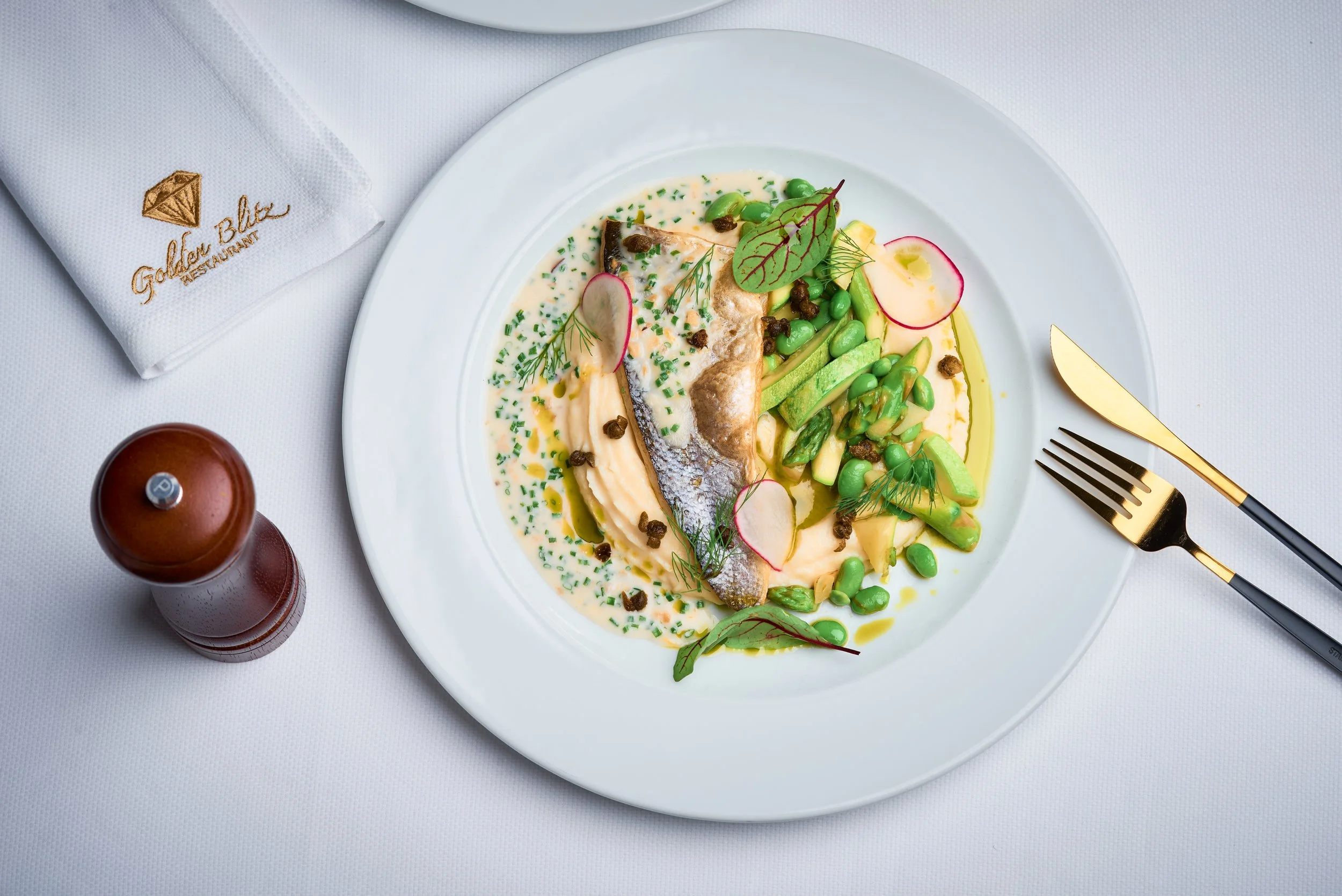 A white plate with a serving of fish fillet topped with sauce and herbs, accompanied by sliced radishes, green beans, and green peas, garnished with fresh greens and chives. The plate is on a white tablecloth with gold-colored cutlery, a white napkin