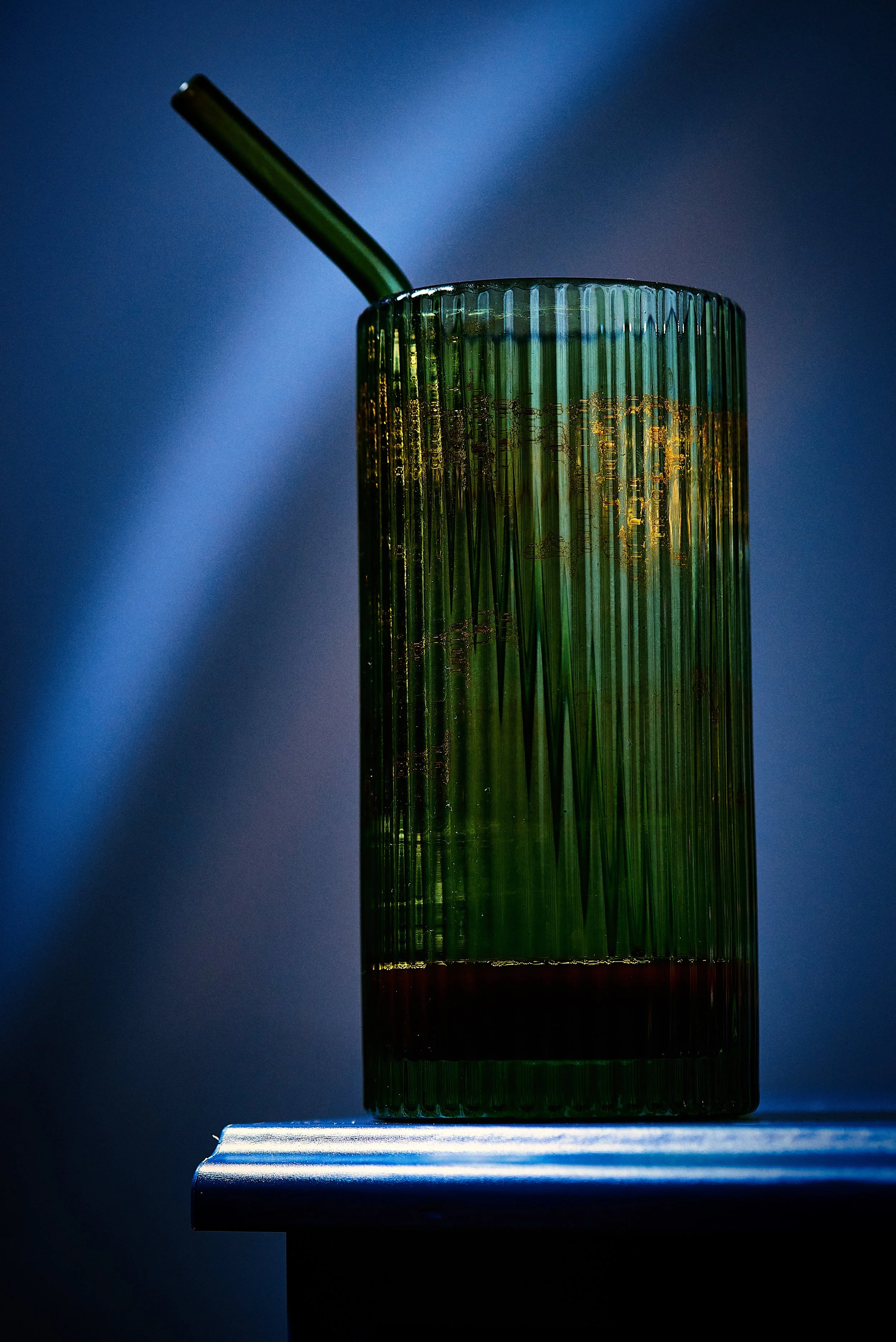 Close-up of a green and amber glass tumbler with vertical ridges, placed on a white surface, against a dark blue background, with a green straw inside.