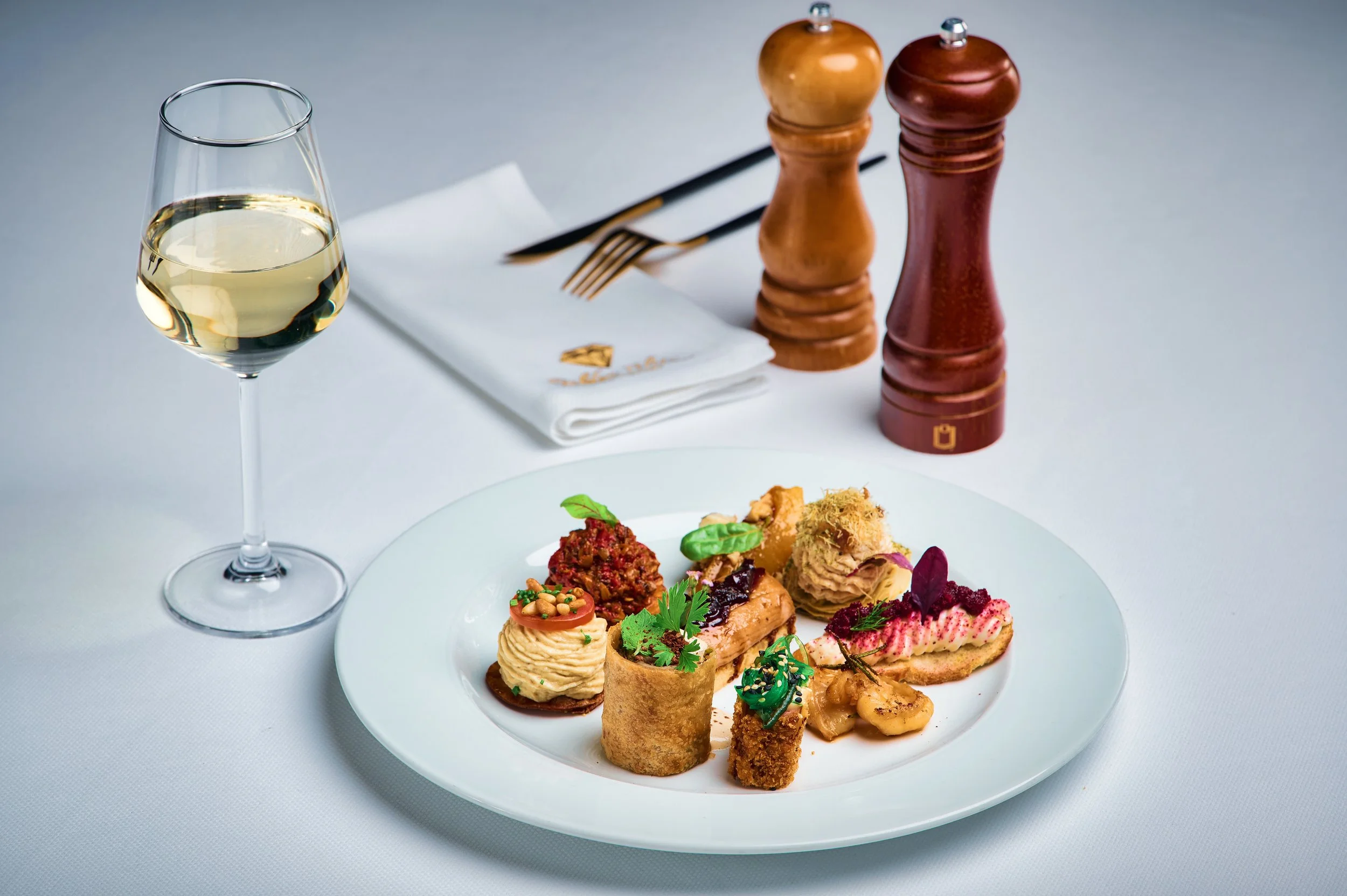 A glass of white wine, a plate of assorted gourmet appetizers including mini tartlets, finger sandwiches, and savory bites, and salt and pepper shakers on a dining table with a soup spoon and napkin in the background.