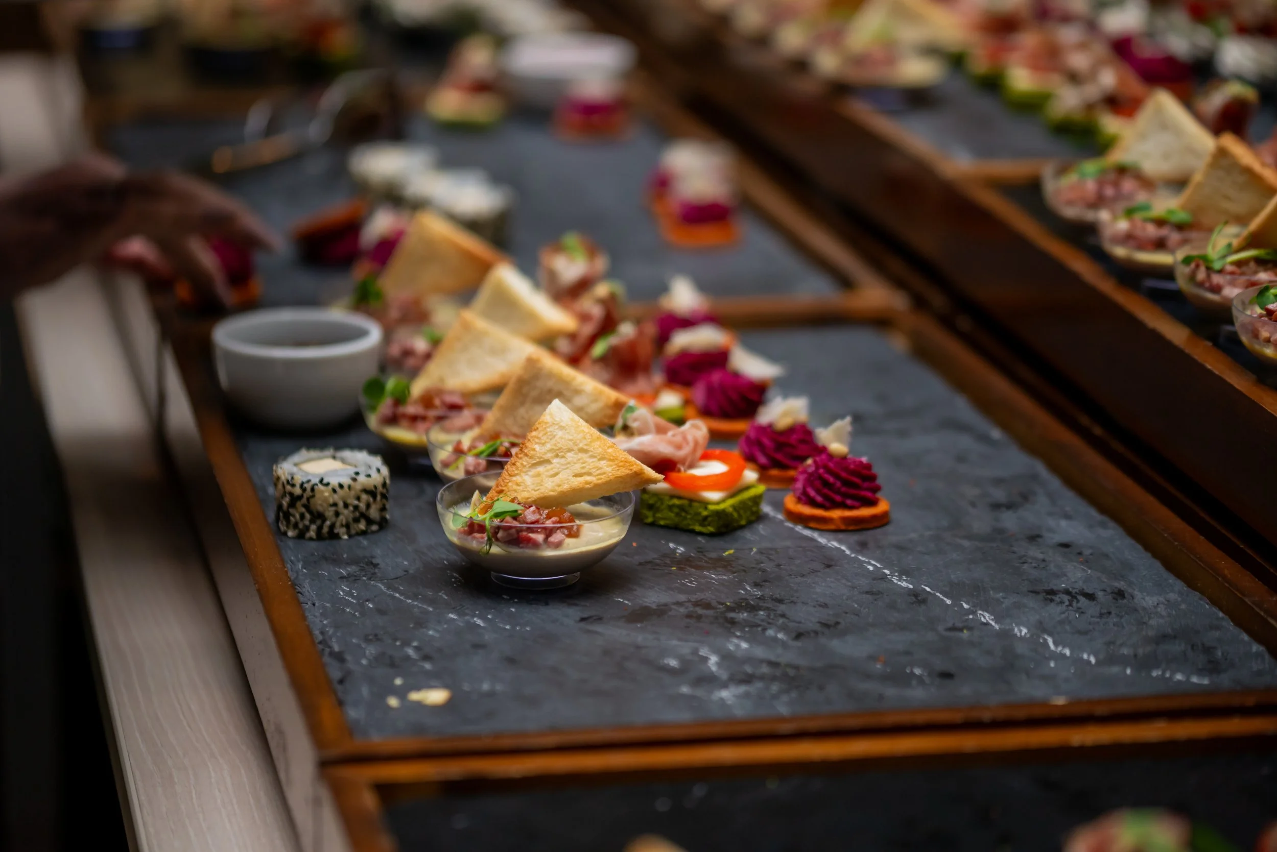 A long table covered with a dark cloth displays various small food appetizers, including sandwiches, sushi, dips, and colorful garnishes, arranged for a gathering or event.