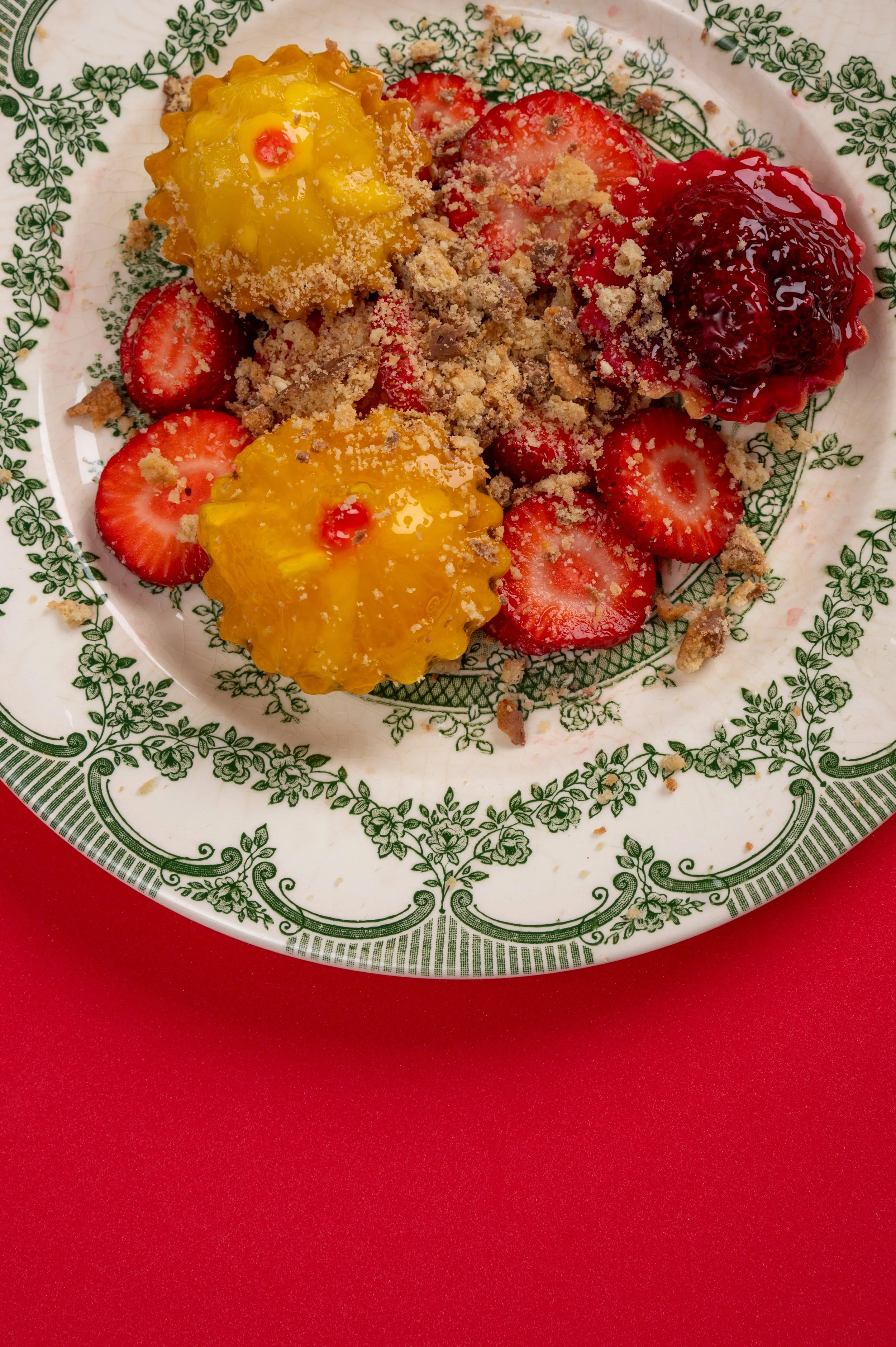 Plate with three cupcakes topped with yellow and red icing and strawberry slices, sprinkled with crushed cookies, on a decorative green and white floral plate, against a red background.