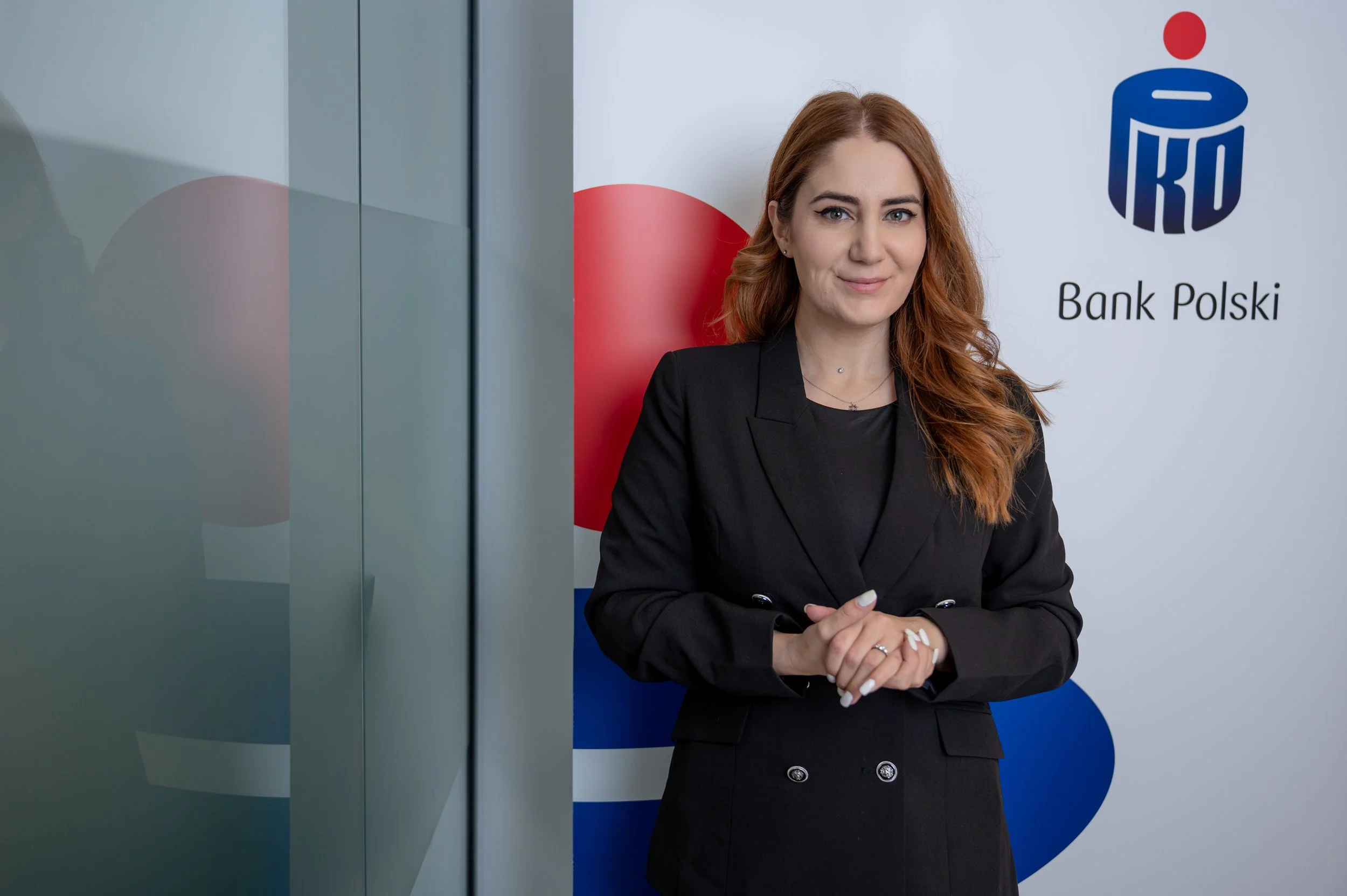 A woman with red hair and a black blazer is standing in front of a backdrop with the logos of Bank Polski. She is smiling subtly and has her hands clasped in front of her.