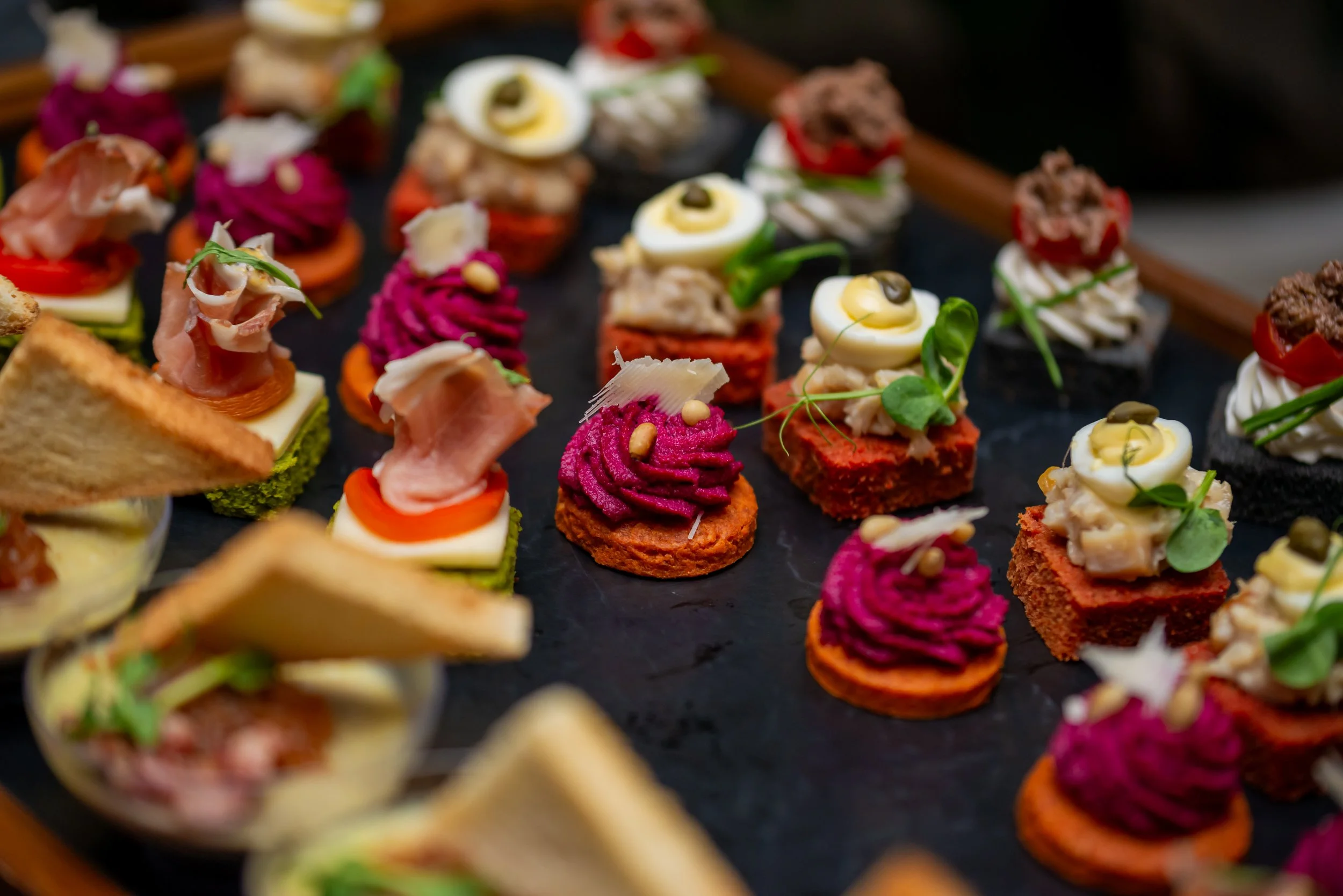 Assorted mini appetizers on a black tray, including topped toast with various spreads, garnishes, and toppings.