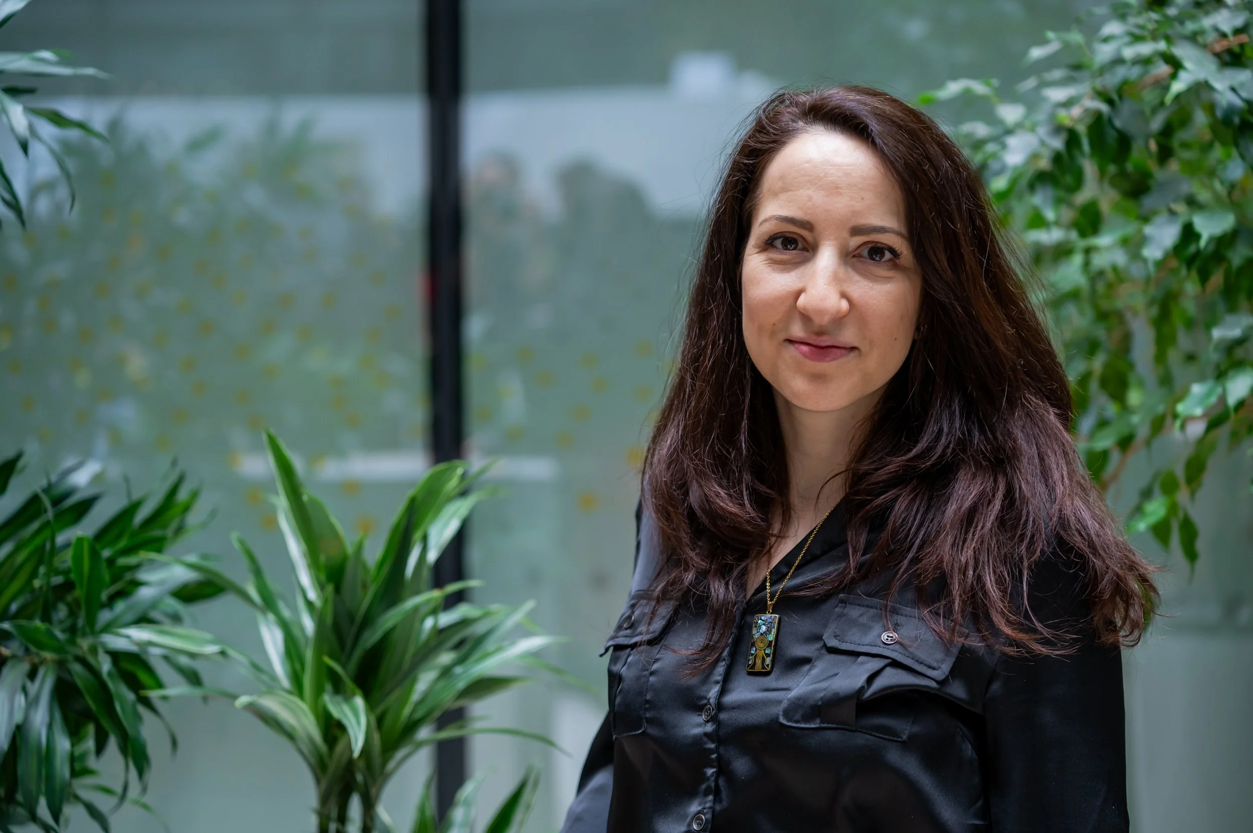A woman with long curly brown hair standing among green plants indoors, wearing a black jacket with a necklace.