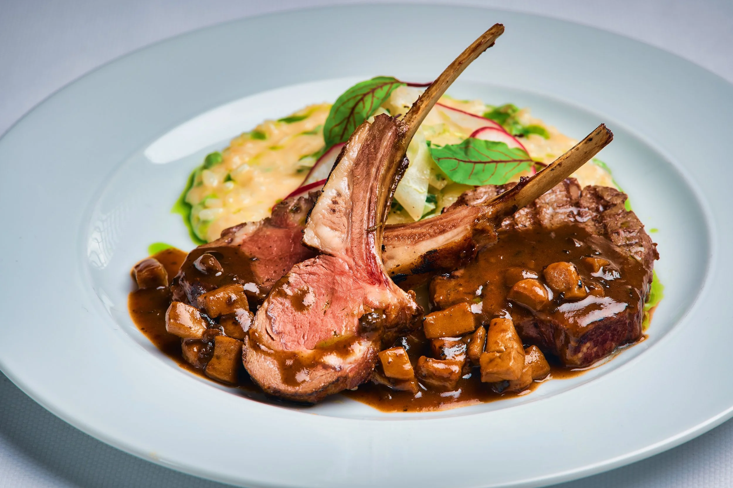 A plated dish featuring a cut of lamb with roasted vegetables, garnished with fresh basil and radish slices, served with a side of creamy mashed potatoes and green sauce.