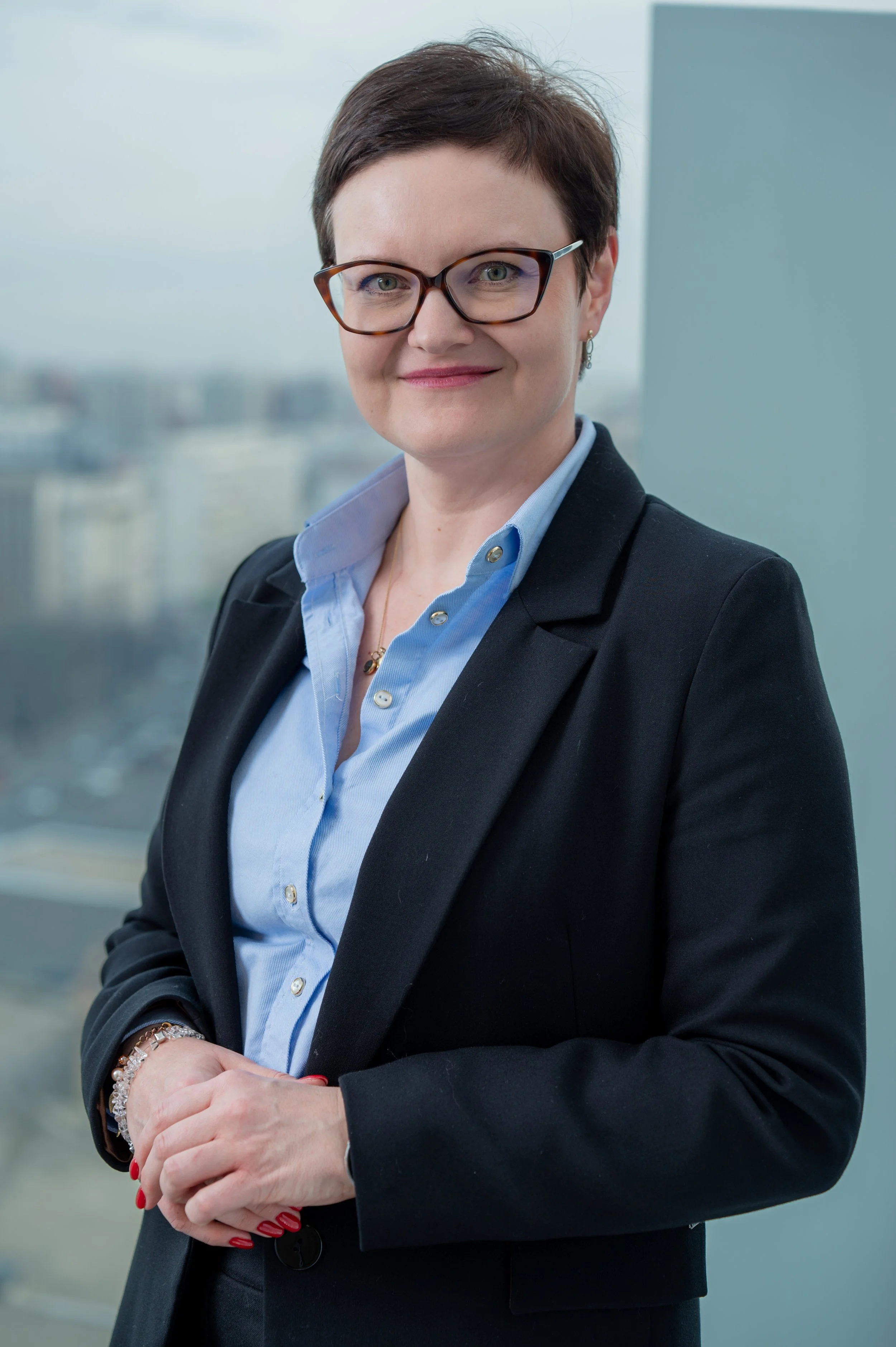 A woman with short dark hair, glasses, and a confident smile, dressed in a blue shirt and black blazer, standing in a modern office with a cityscape background.