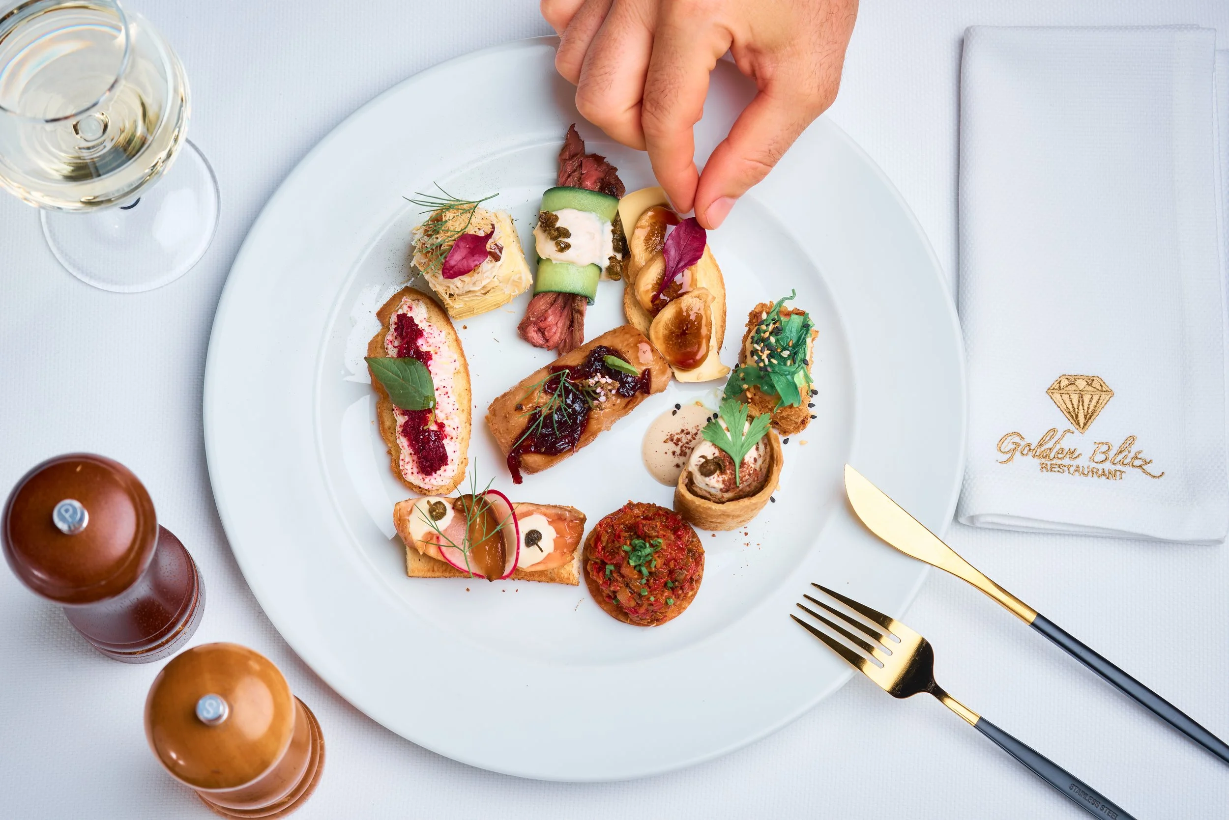 A white plate with various appetizers, a hand pinching food, a glass of white wine, and a set of gold utensils on a white tablecloth with a napkin embroidered with a diamond logo.