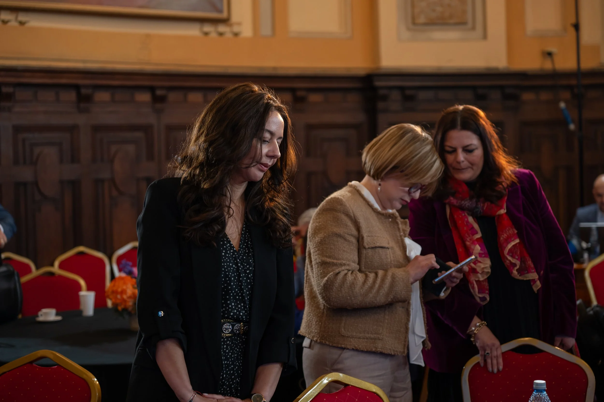 Three women standing with heads bowed in prayer or reflection in a conference room with red chairs and dark wood paneling.