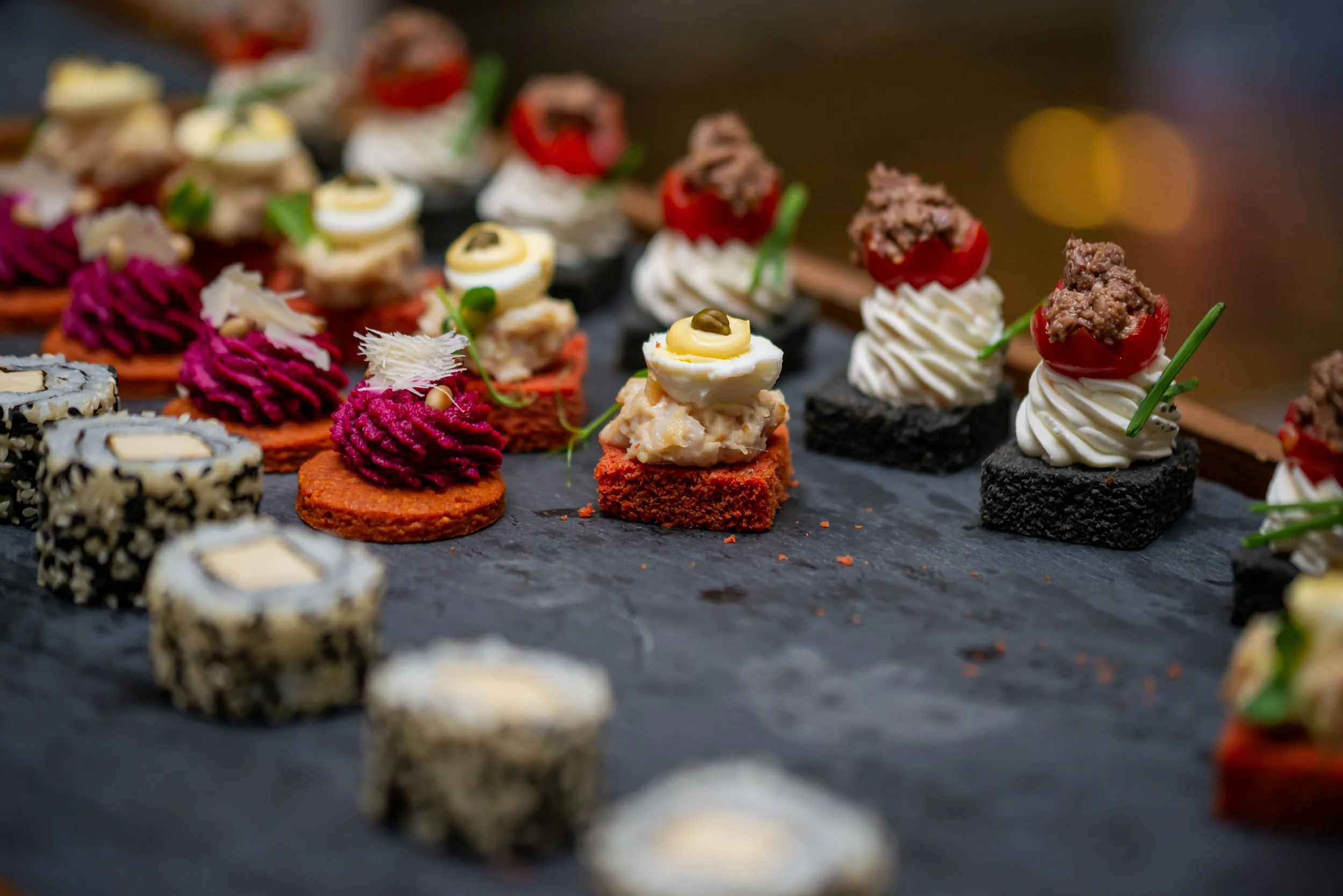 Assorted colorful appetizers or canapés arranged on a dark serving platter, featuring various toppings and garnishes.