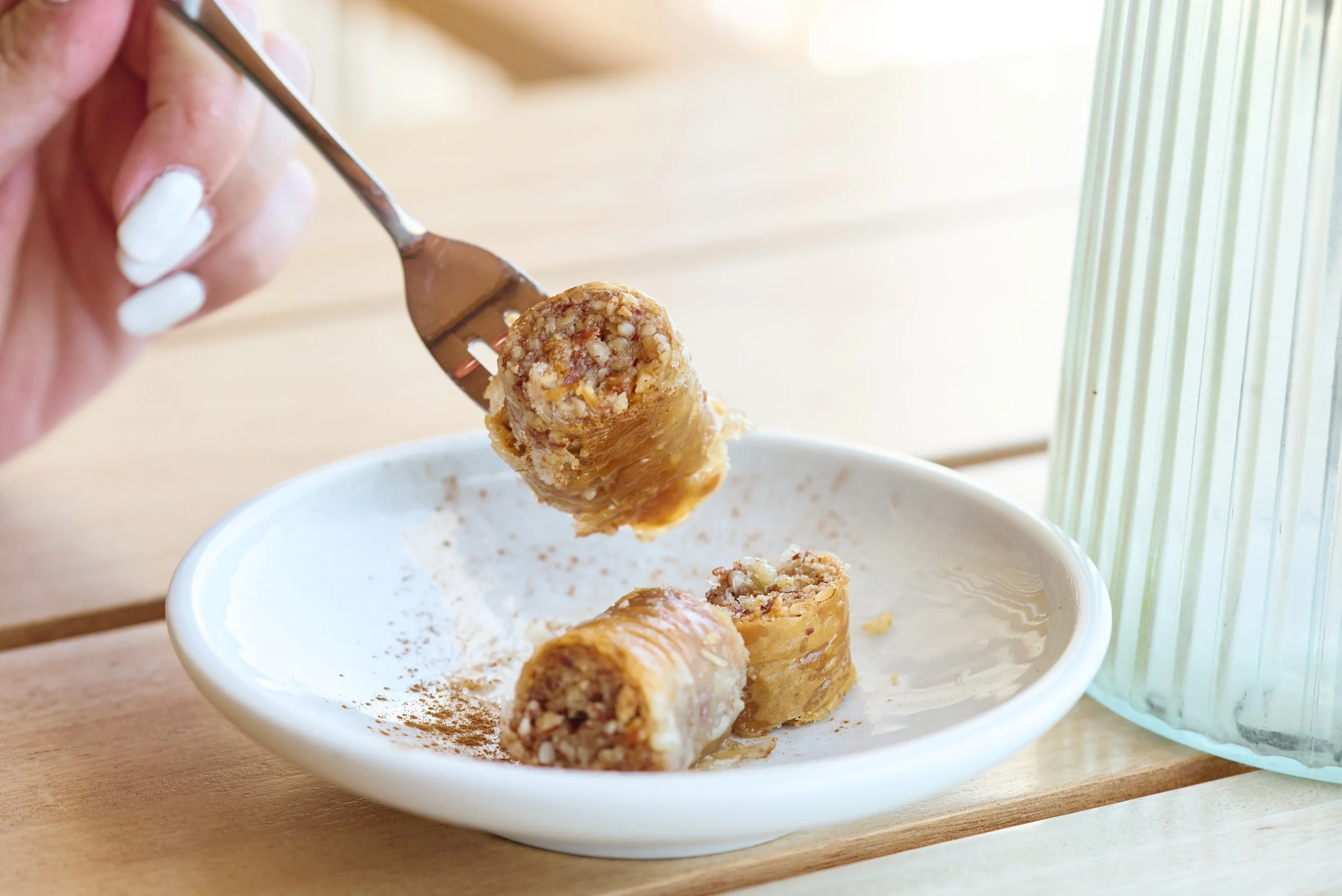 Close-up of a hand holding a spoon with a peanut butter rice crispy treat over a white bowl on a wooden surface, with a glass of water and window in the background.