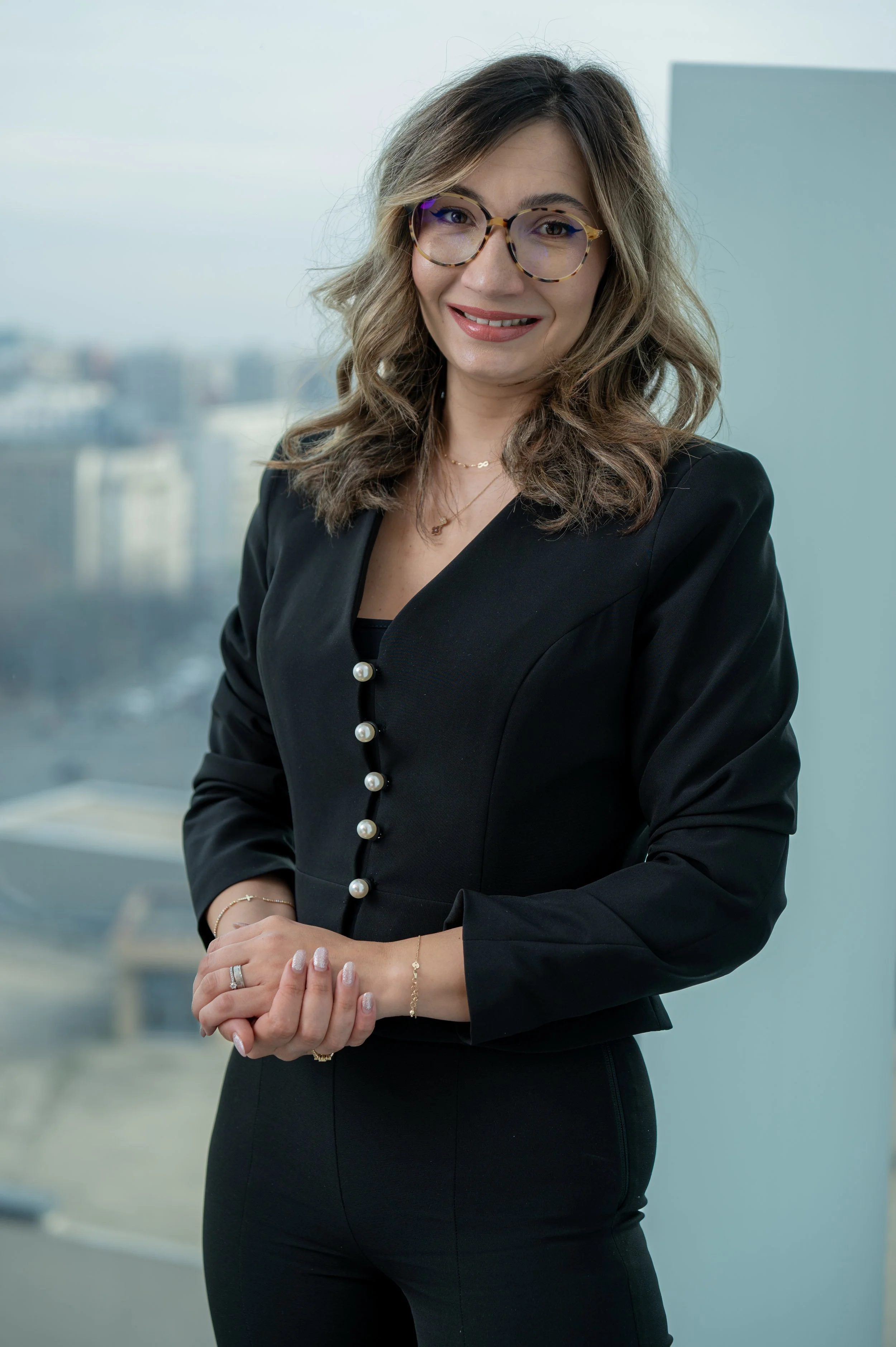 Professional woman in black blazer with pearl buttons, glasses, and jewelry, standing in an office with cityscape view through window.