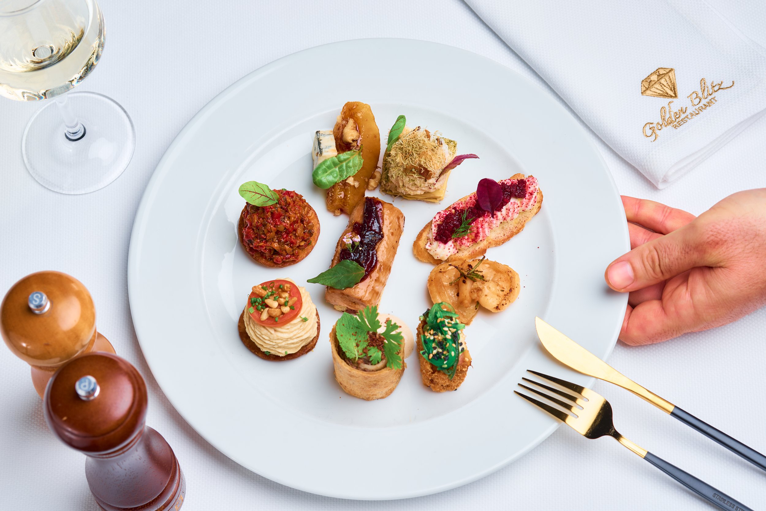 Assorted small gourmet appetizers and desserts on a white plate, with a glass of white wine nearby. The plate includes canapés topped with various garnishes, and the setting suggests a fine dining experience at the Golden Bluff Restaurant.
