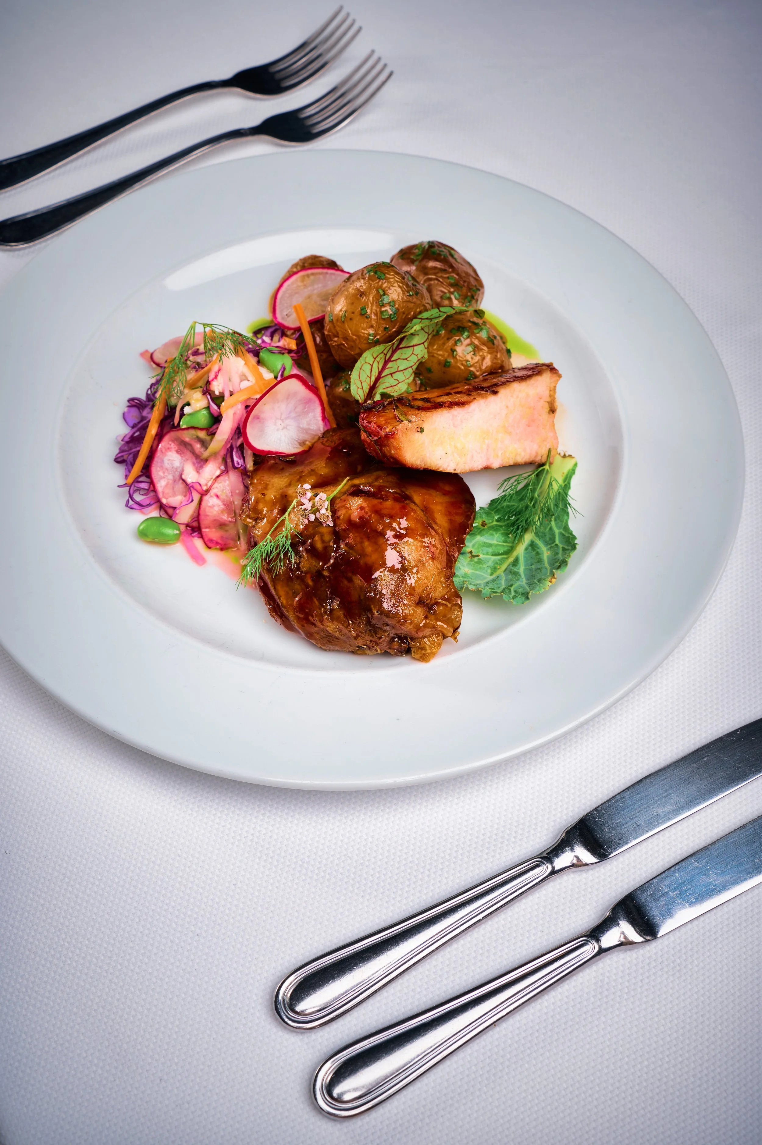 A white plate of gourmet food featuring glazed potatoes, grilled meat, salad with radish slices, and herbs. The plate is on a white tablecloth with a fork and knife beside it.
