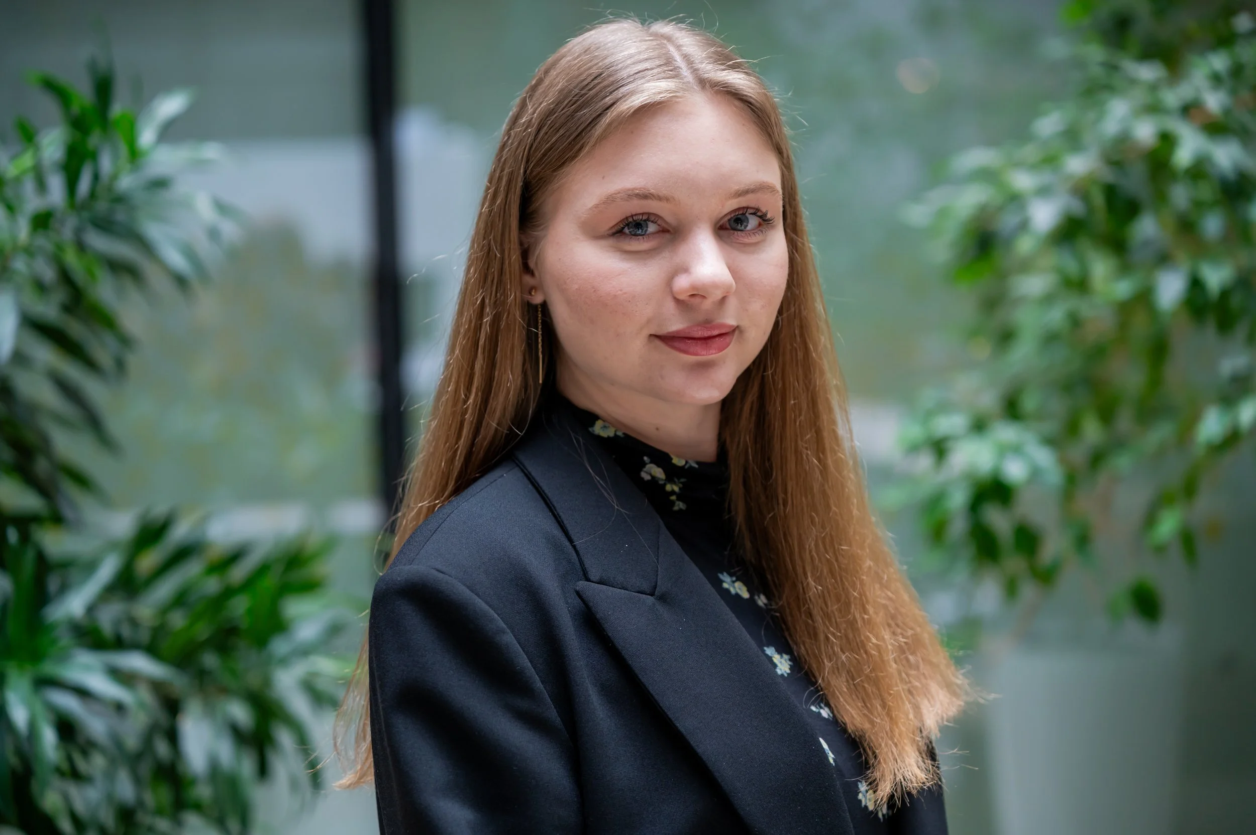 A young woman with long red hair, light skin, and blue eyes, wearing a dark blazer and a black shirt with small floral patterns, standing outdoors near green foliage.