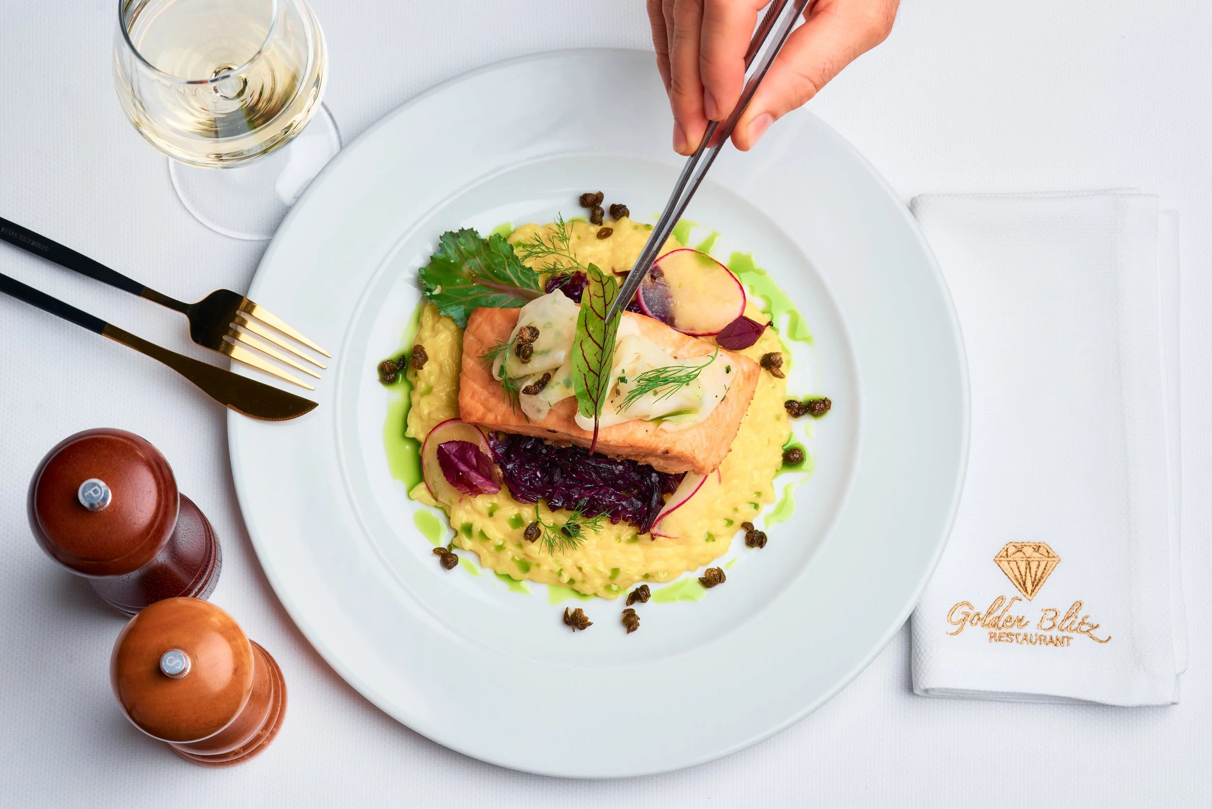 A white plate of salmon with sauces, herbs, and garnishes, accompanied by a glass of white wine on a white tablecloth.