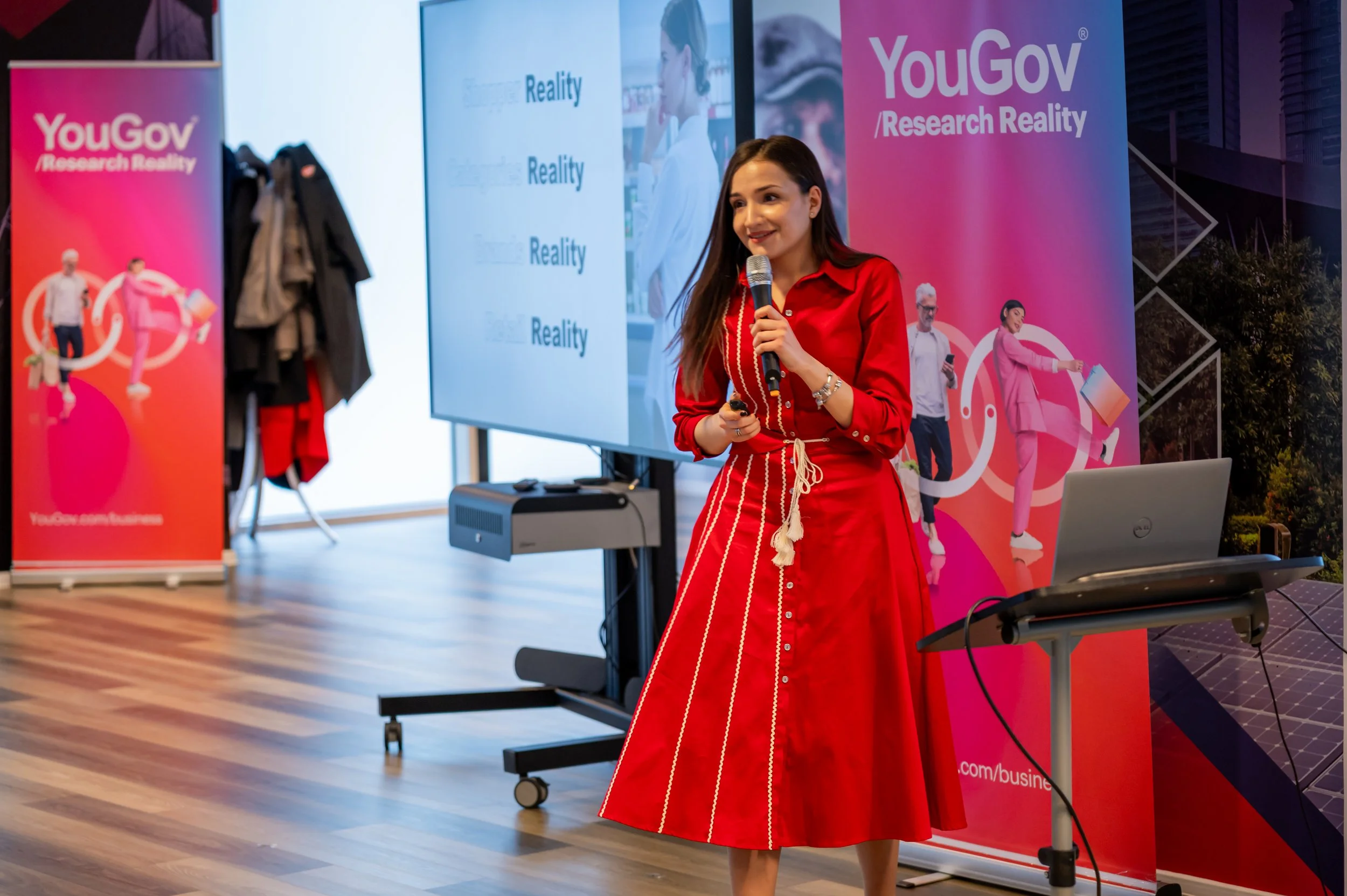 A woman in a red dress holding a microphone presents in front of banners that read 'YouGov Research Reality' at a conference or event.