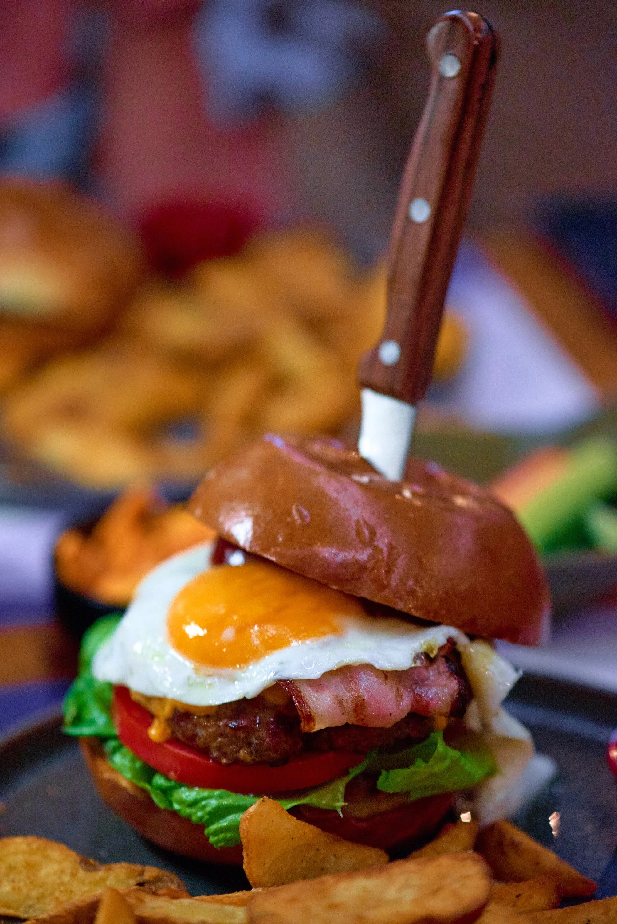 A close-up of a gourmet burger with a fried egg, bacon, tomato, lettuce, and a brioche bun, served with potato chips.
