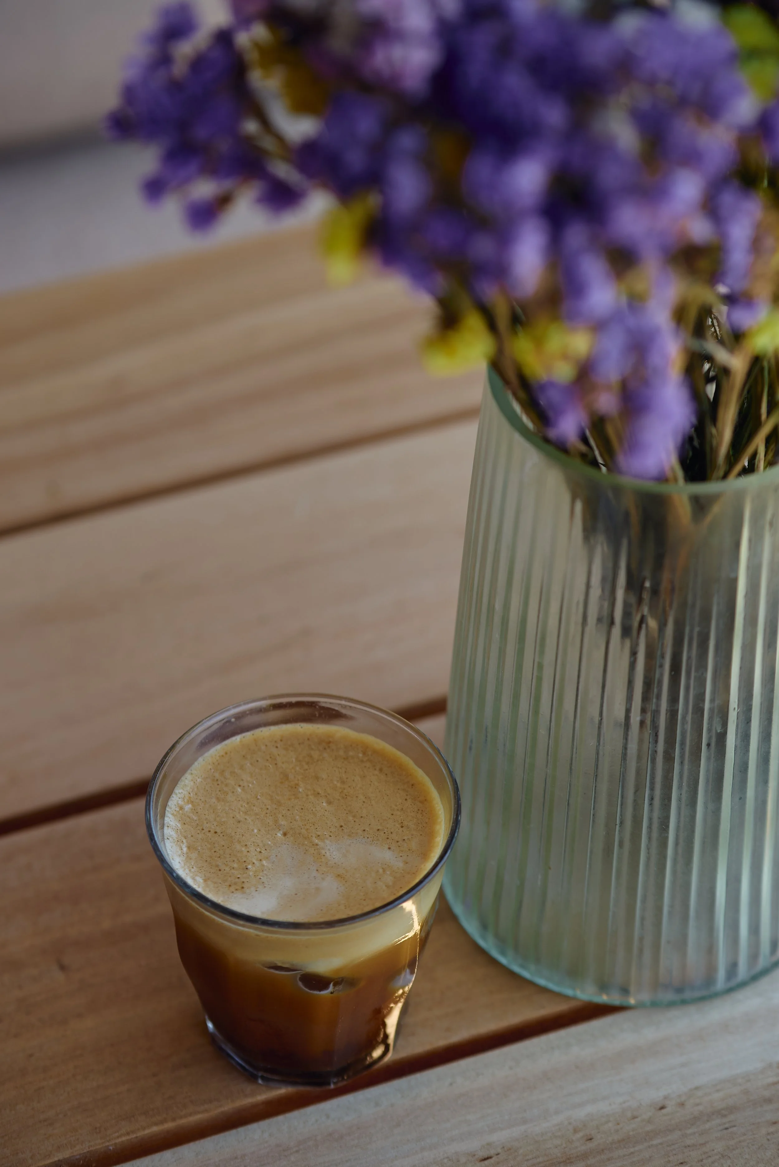 A glass of coffee with a foam top, placed on a wooden surface. In the background, a vase with purple flowers is partially visible, sitting on the same wooden surface.