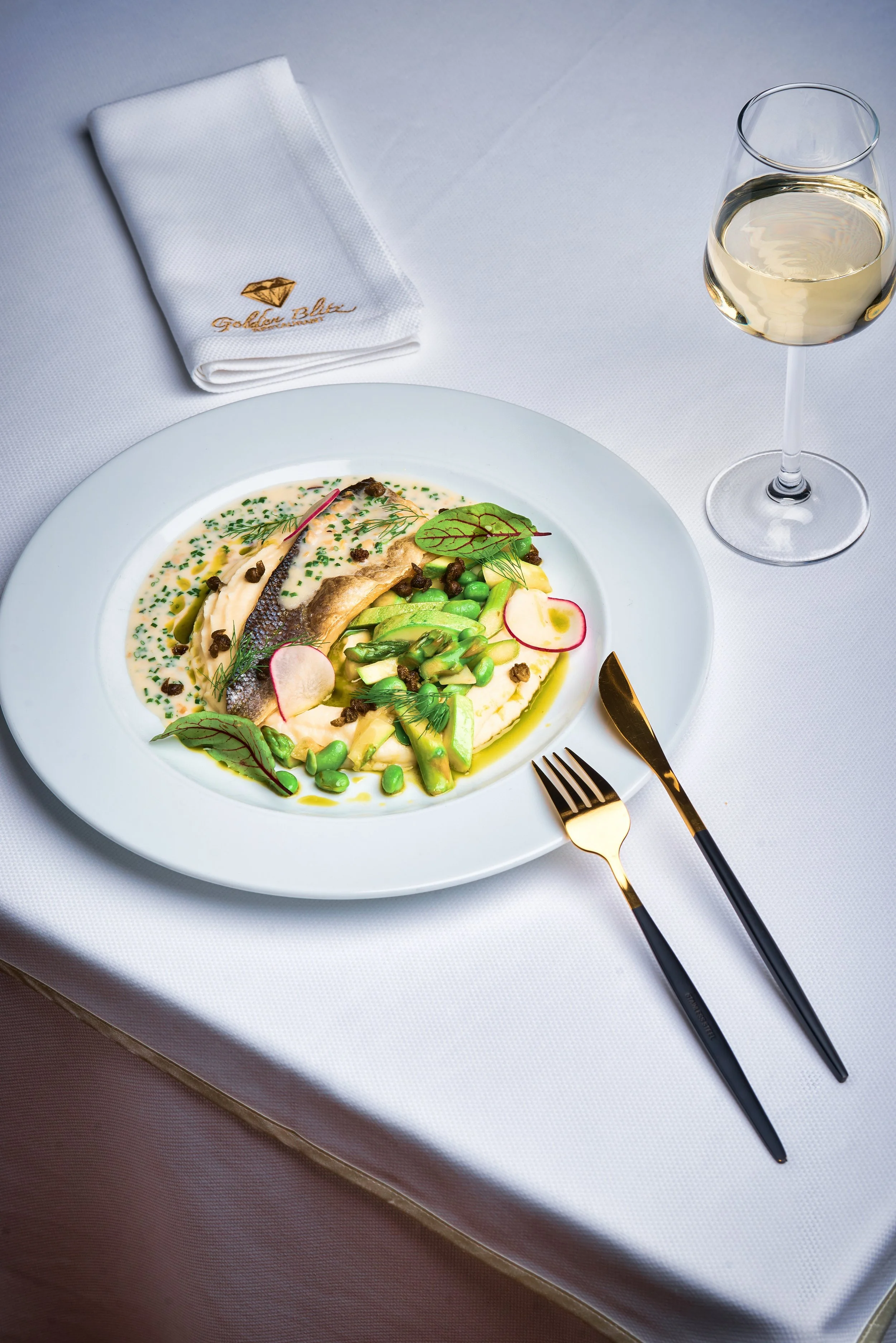 A white dinner plate with fish, green vegetables, radish slices, and herbs, accompanied by a glass of white wine, set on a white tablecloth with a folded napkin embroidered with a gold diamond and the words "Golden Blue".