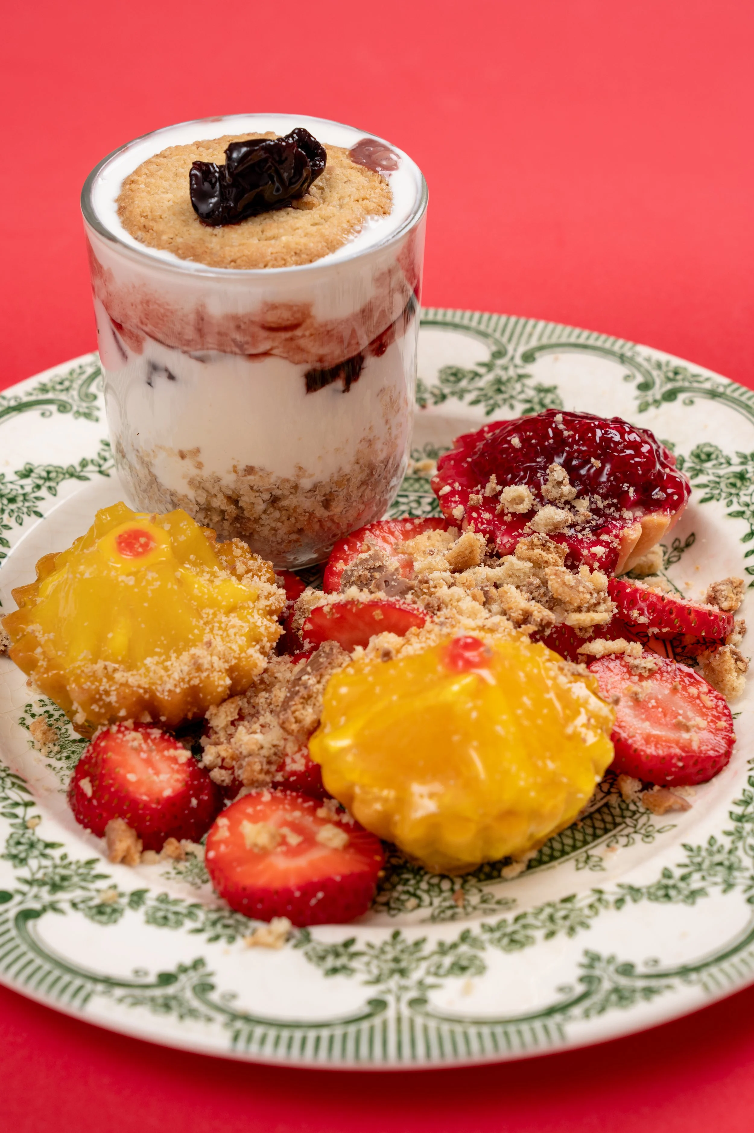 A dessert with strawberries, yellow custard, and crumbled topping on a decorative plate, with a layered parfait in a glass
