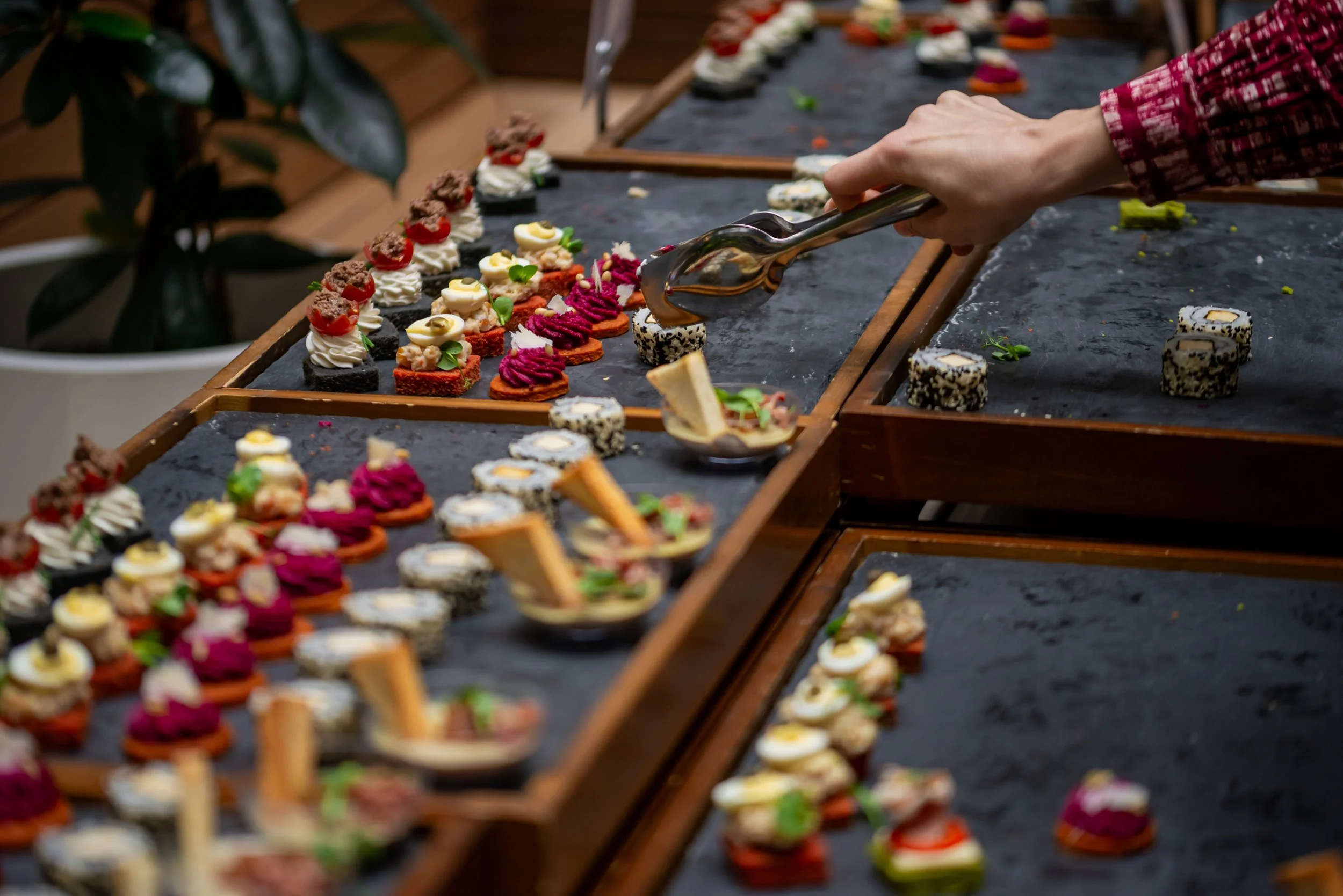 A person using tongs to arrange a variety of colorful, bite-sized appetizers and sushi on a black tray at a catering or buffet setup.