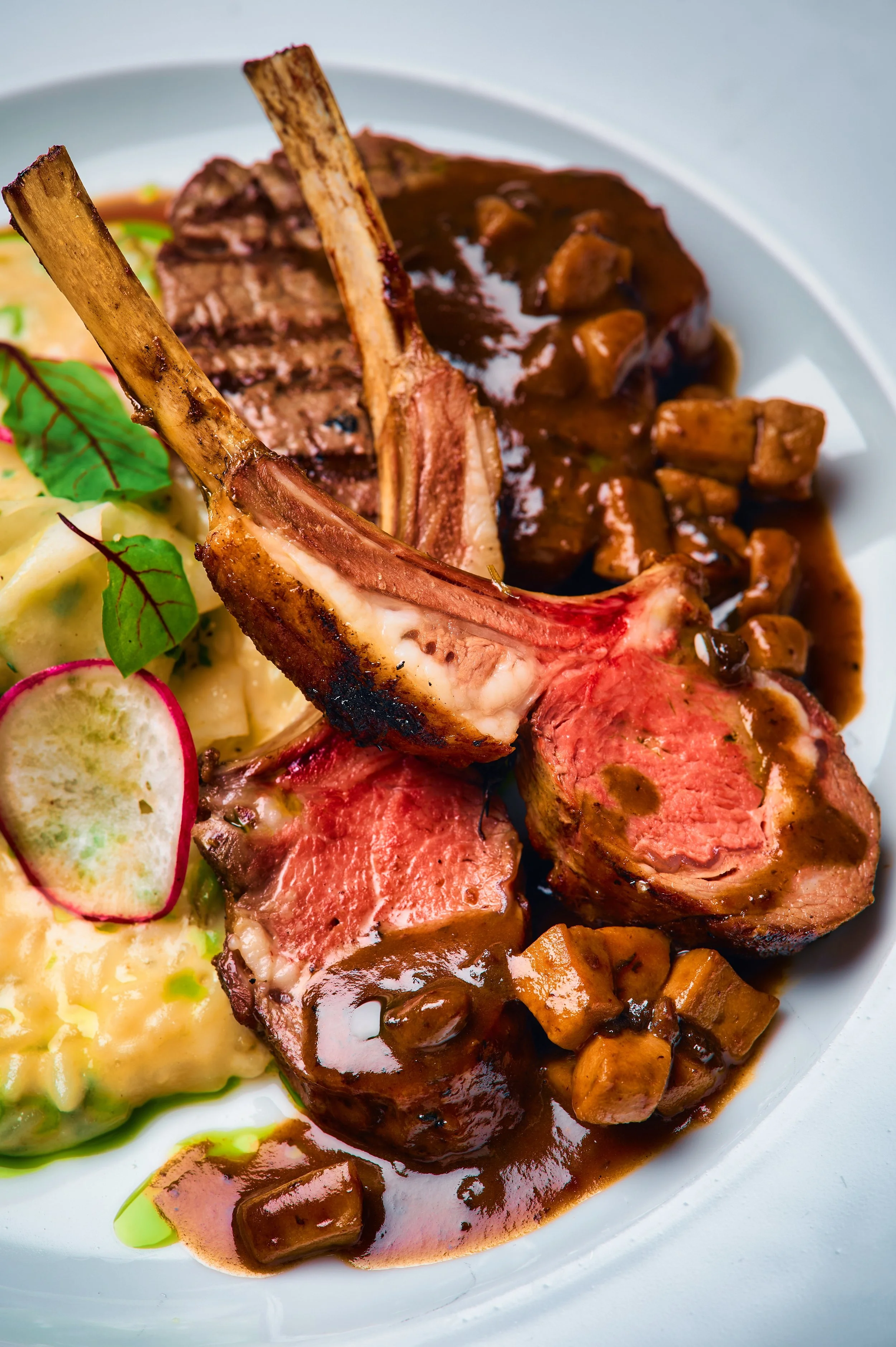 Close-up of a plate featuring a cooked lamb chop with a bone, smothered in a brown gravy with mushrooms, alongside mashed potatoes garnished with radish slices and herbs.